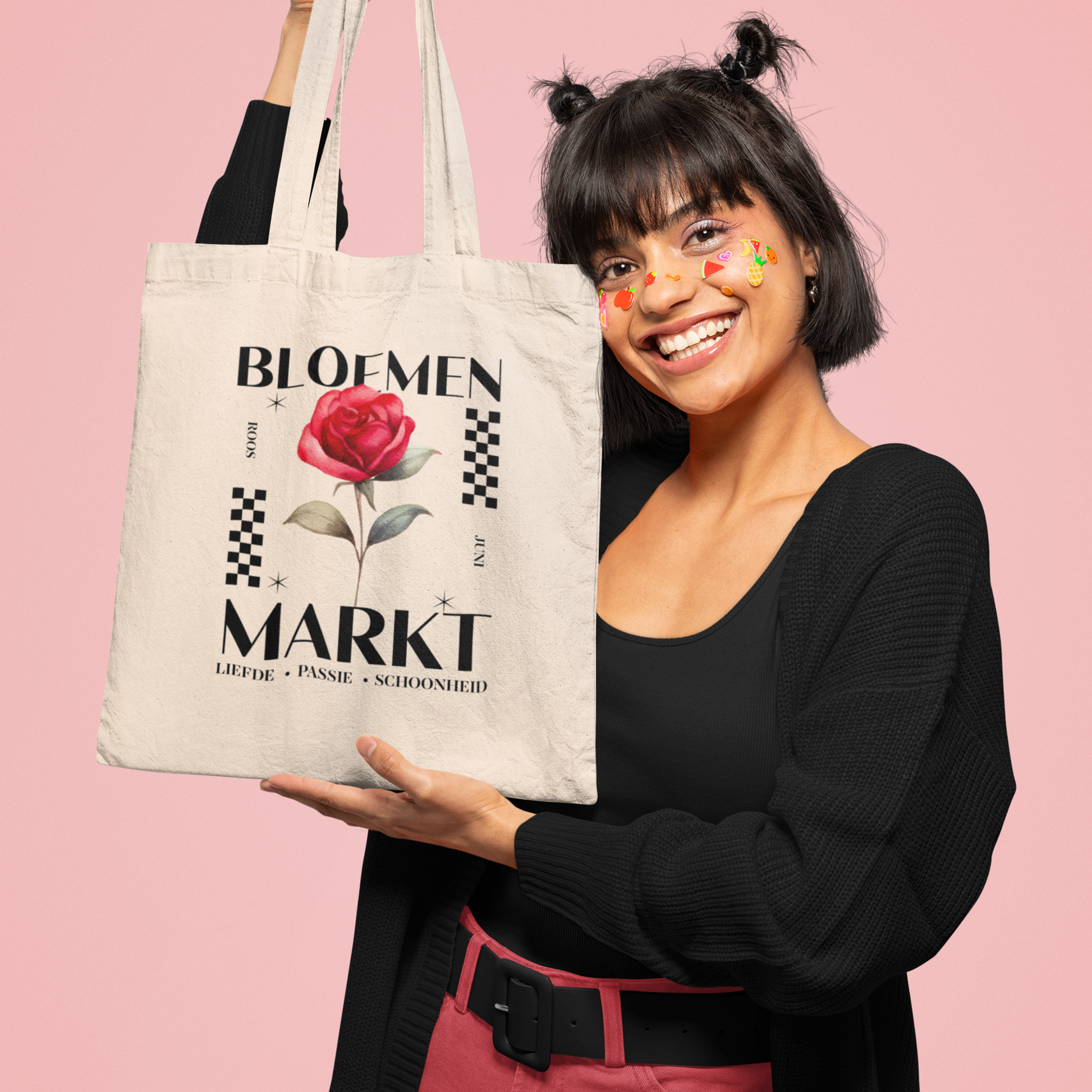 misje met zwart haar met staartjes en stickers op haar gezicht die een tote bag vast houdt,totebag met  de tekst bloemenmarkt en een illustratie van een geboortebloem van de maand juni, een roos. Met onderaan de kernwoorden van deze maand : liefde,passie,schoonheid