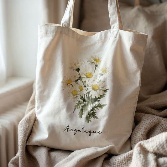 Canvas totebag met madeliefjes en naam Angelique, liggend op een zacht plaid bij een raam.