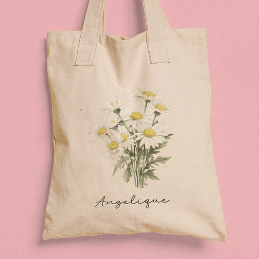 Gepersonaliseerde totebag met madeliefjes en de naam Angelique, gefotografeerd tegen een roze achtergrond.