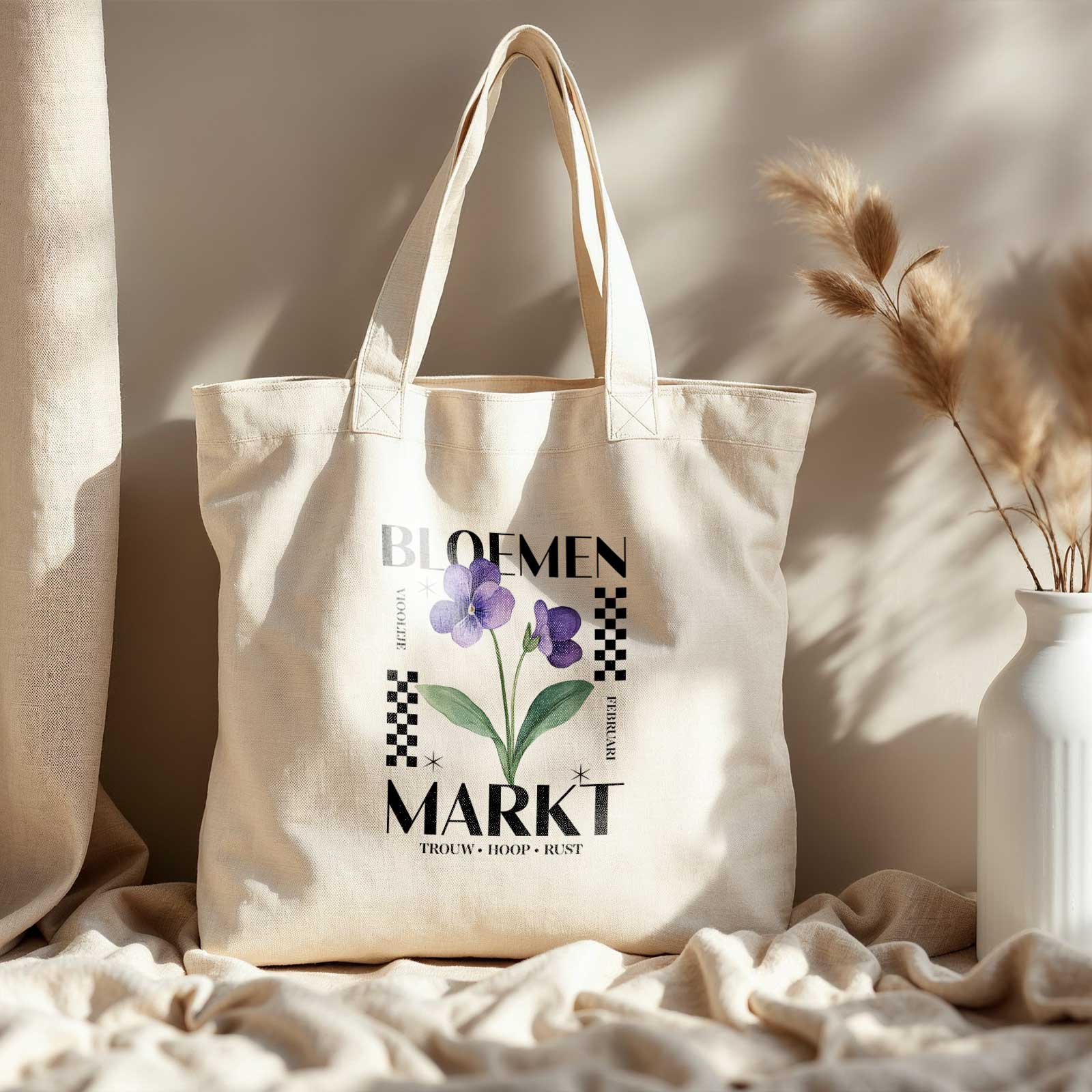 totebag met  de tekst bloemenmarkt en een illustratie van een geboortebloem van de maand februari, een viooltje. Met onderaan de kernwoorden van deze maand : trouw, hoop, rust. Totebag staat in het licht op een plek met droogbloemen en een creme kleed