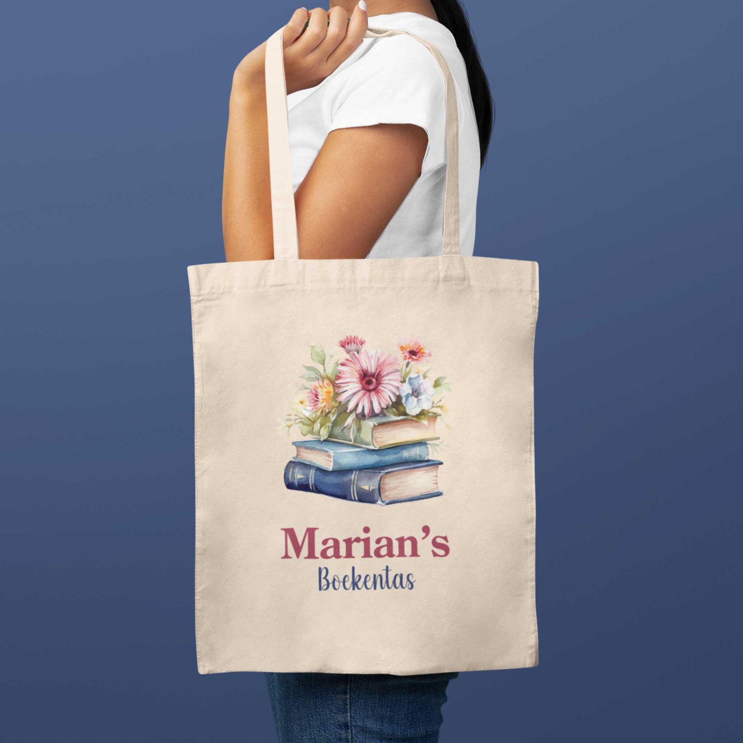Vrouw draagt een crèmekleurige totebag met bloemen en boeken illustratie en de tekst "Marian's Boekentas", tegen een blauwe achtergrond