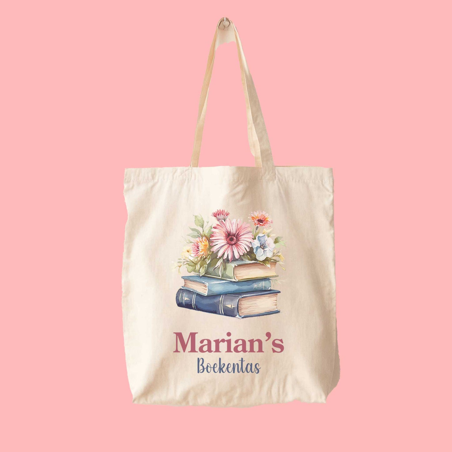 Crèmekleurige katoenen totebag met boekillustratie en kleurrijke bloemen, voorzien van de opdruk "Marian's Boekentas", op een roze achtergrond