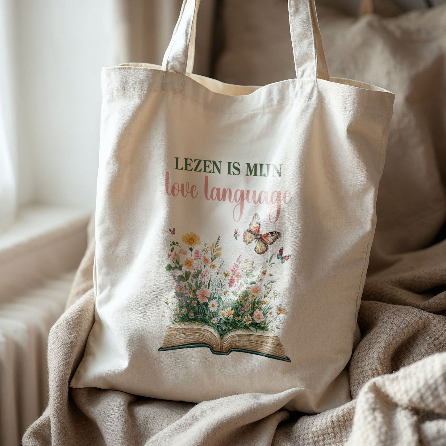 Katoenen totebag met illustratie van open boek, bloemen en vlinders en tekst Lezen is mijn love language, liggend op plaid