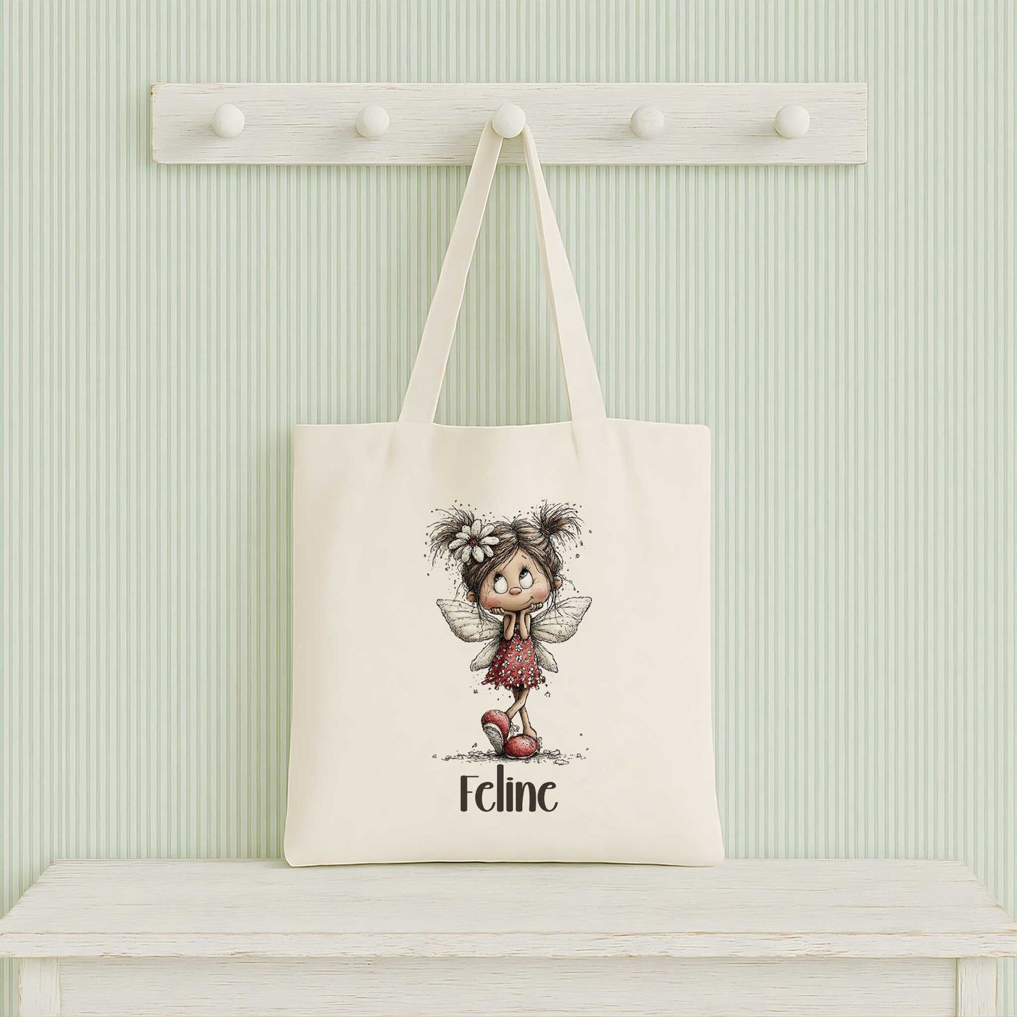 Beige totebag hangend aan een witte kapstok tegen een lichtgroene gestreepte muur, bedrukt met een fee en de naam Feline.