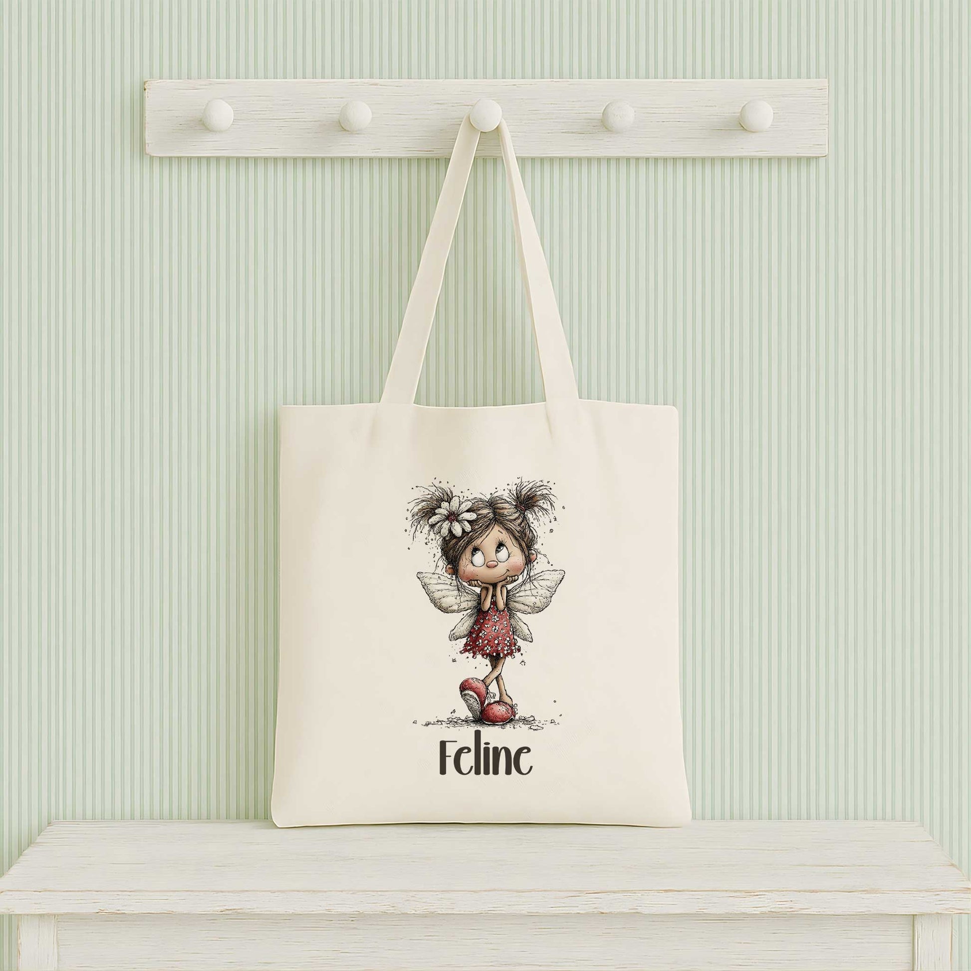 Beige totebag hangend aan een witte kapstok tegen een lichtgroene gestreepte muur, bedrukt met een fee en de naam Feline.