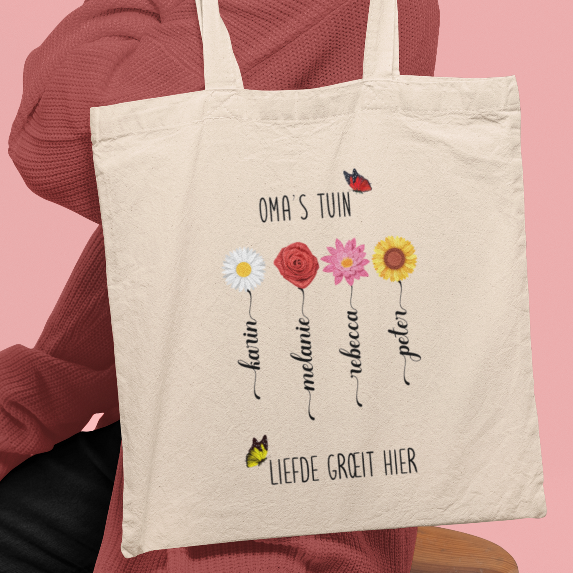 Totebag ‘Oma’s Tuin’ gedragen over schouder, vier bloemen met namen zichtbaar, roze achtergrond.
