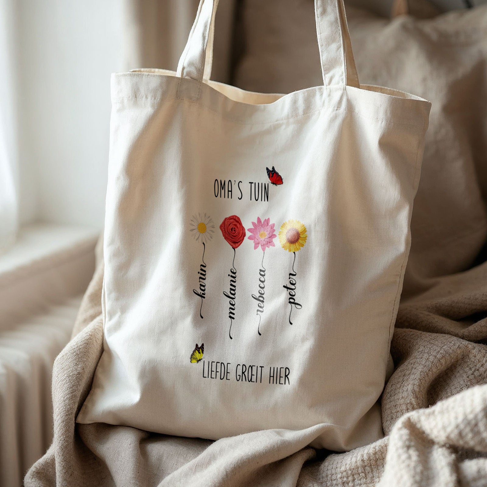 Totebag ‘Oma’s Tuin’ met bloemen en namen op bank bij raam, naturel katoen.