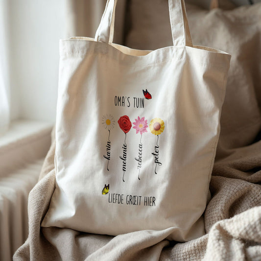 Totebag ‘Oma’s Tuin’ met bloemen en namen op bank bij raam, naturel katoen.