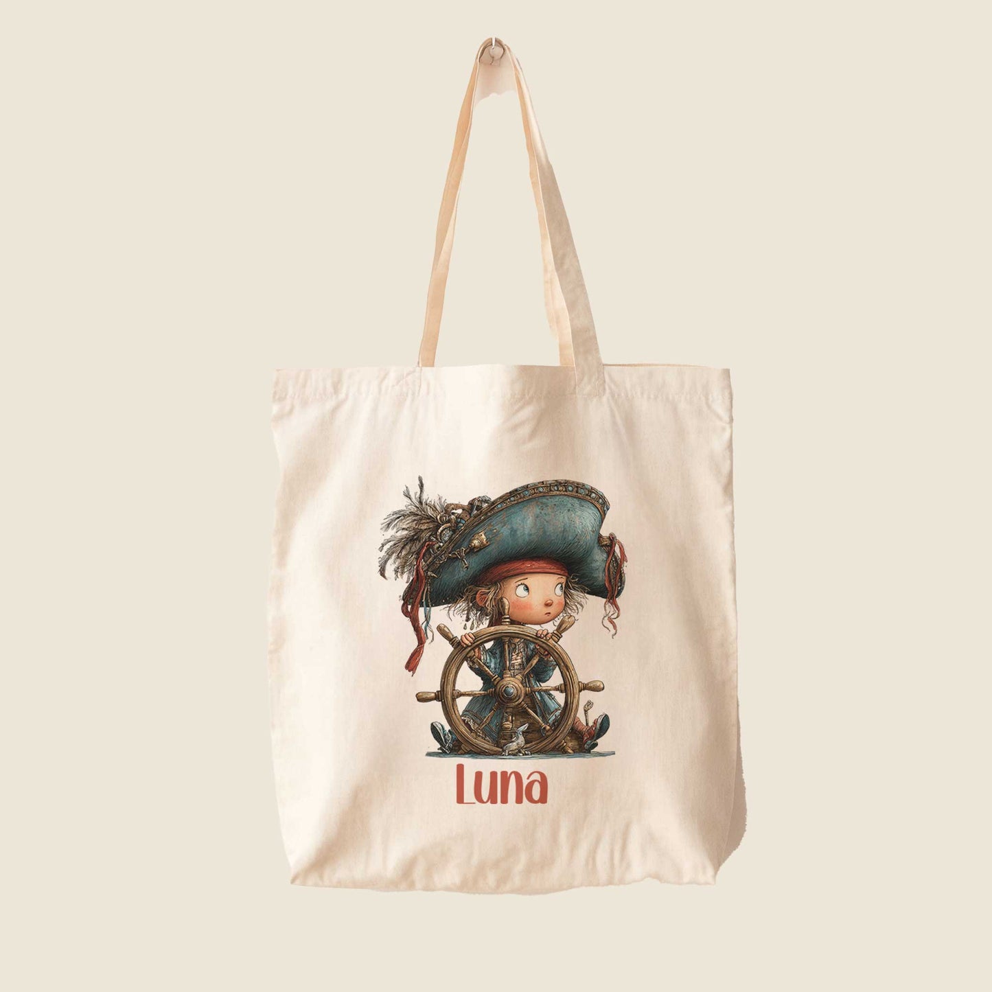 Crèmekleurige totebag op een lichte achtergrond met een illustratie van een piratenkind achter een houten stuurwiel en de naam Luna onder de afbeelding.