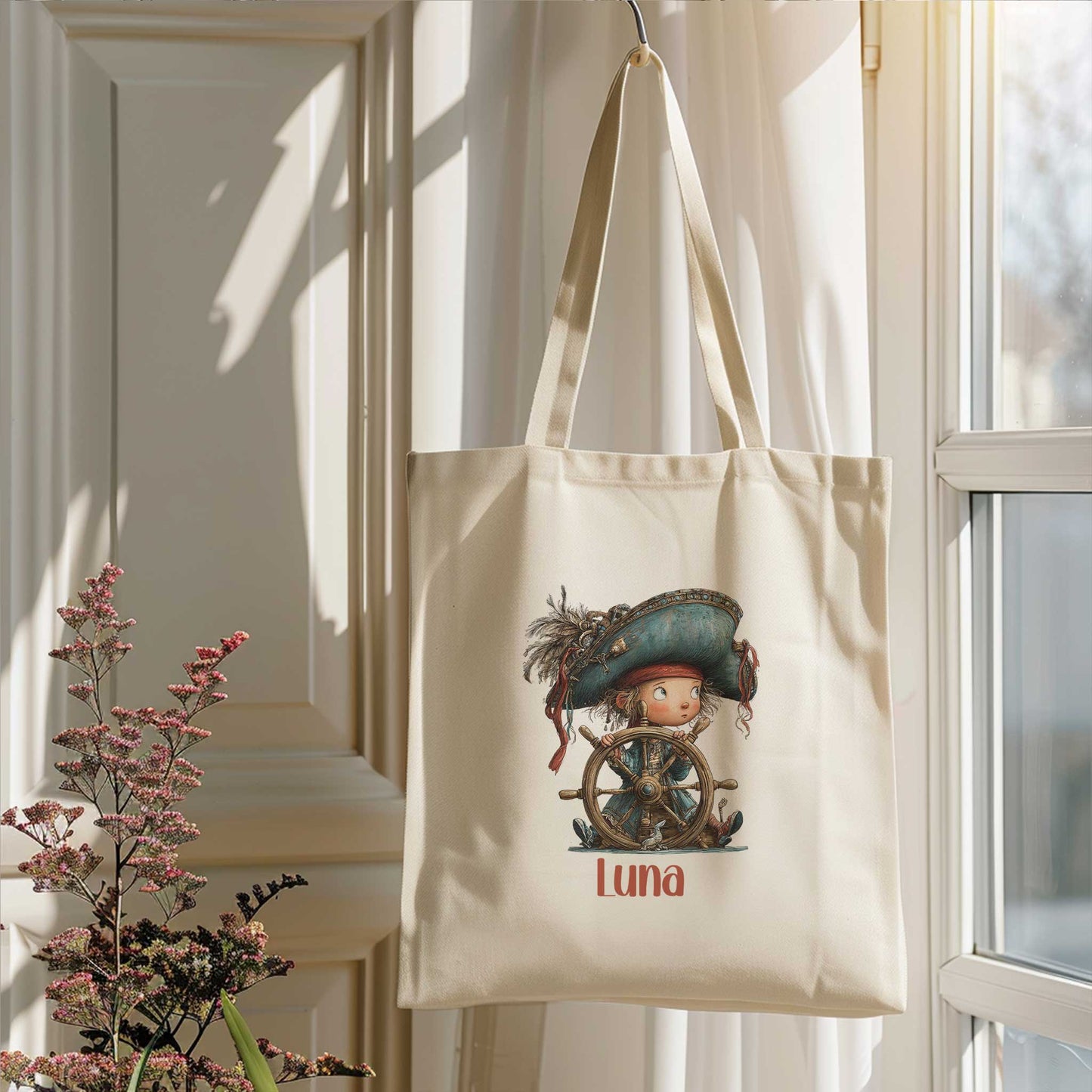 Crèmekleurige totebag hangt aan een haak bij een raam. Op de tas staat een illustratie van een klein piratenkind achter een houten stuurwiel met daaronder de naam Luna.
