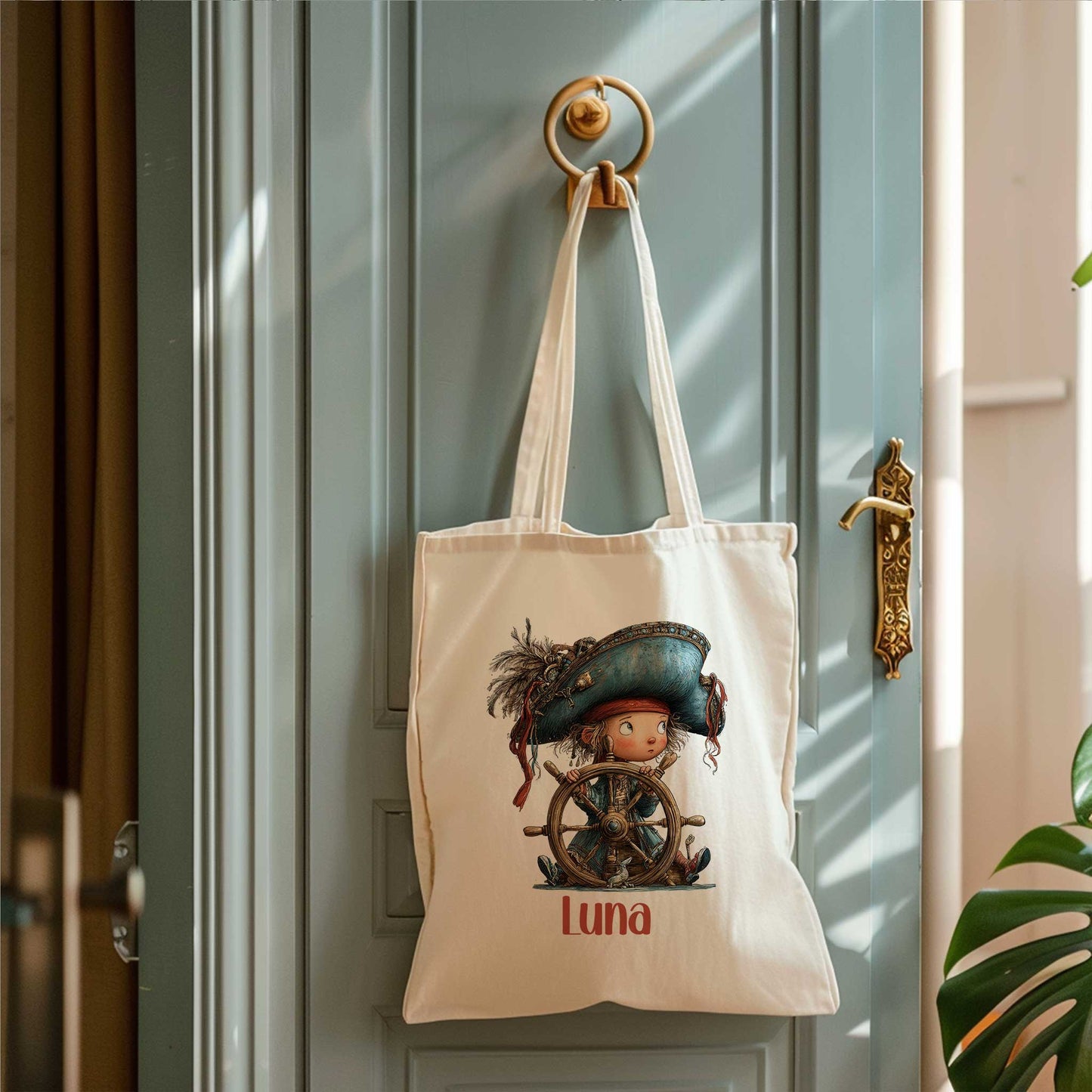 Crèmekleurige totebag hangt aan een ronde wandhaak op een blauwgroene deur. Op de tas staat een piratenkind achter een houten stuurwiel met de naam Luna.