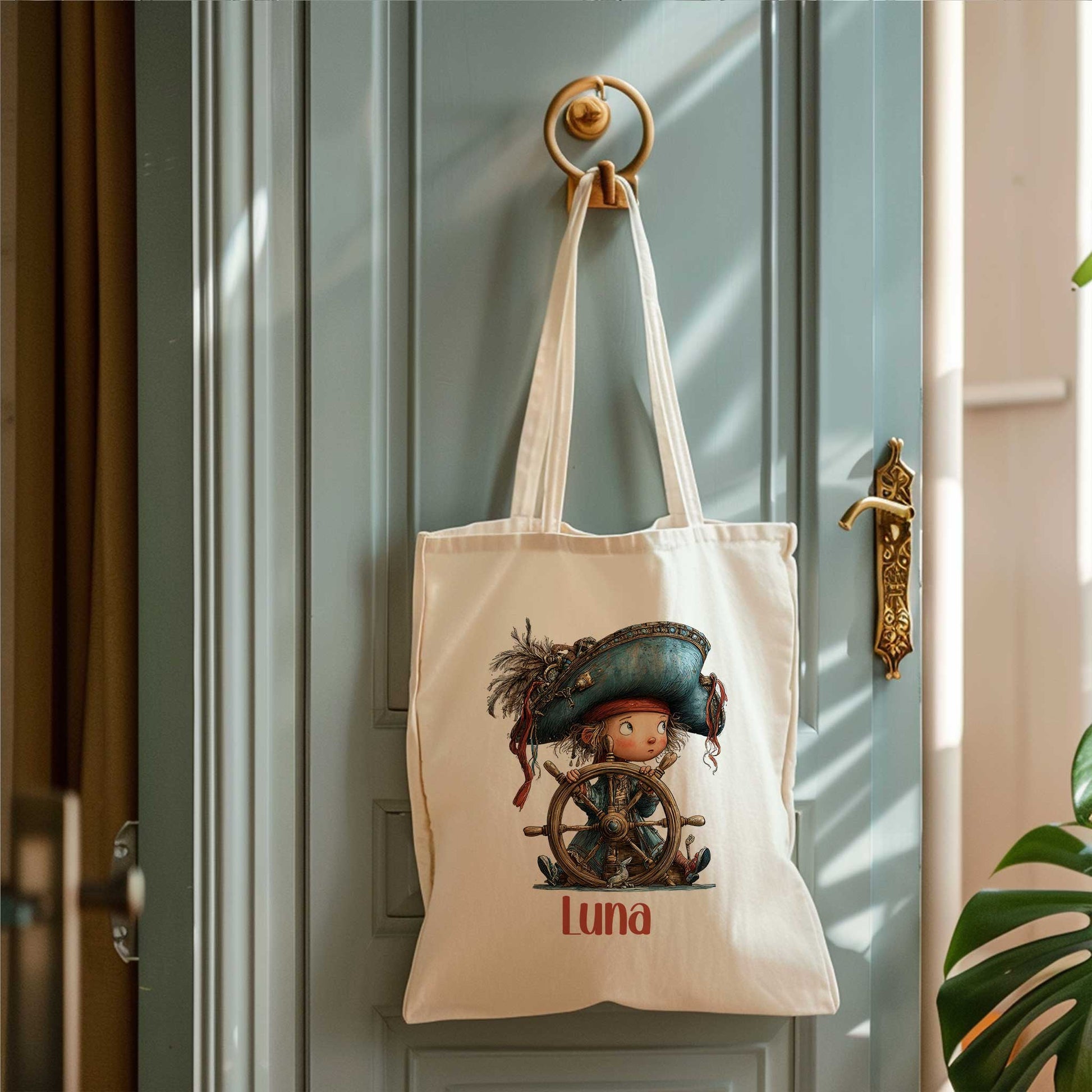 Crèmekleurige totebag hangt aan een ronde wandhaak op een blauwgroene deur. Op de tas staat een piratenkind achter een houten stuurwiel met de naam Luna.