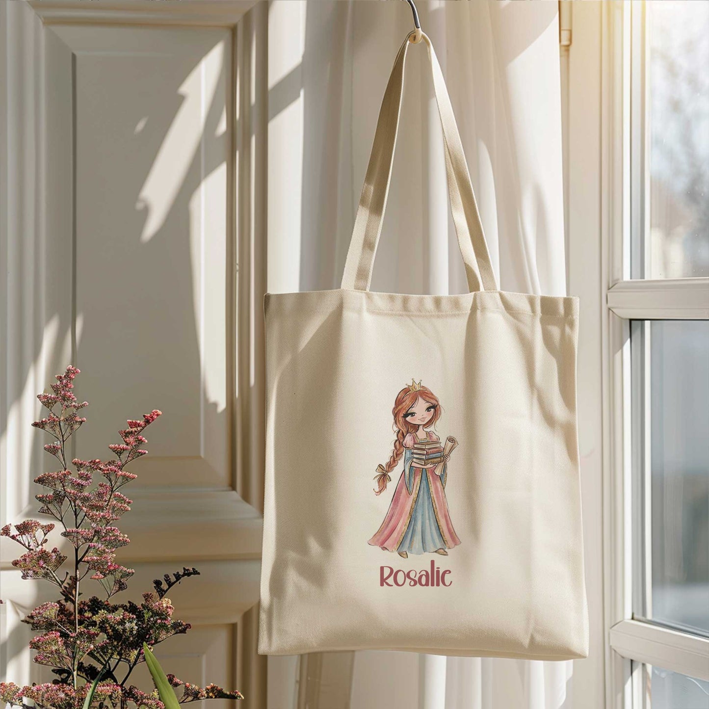 Beige totebag hangend naast een raam in zacht zonlicht. Op de tas staat een prinses met een stapel boeken en de naam Rosalie.