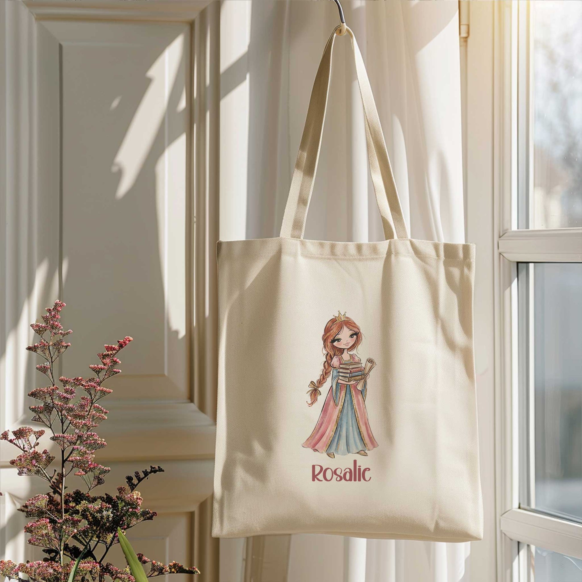 Beige totebag hangend naast een raam in zacht zonlicht. Op de tas staat een prinses met een stapel boeken en de naam Rosalie.