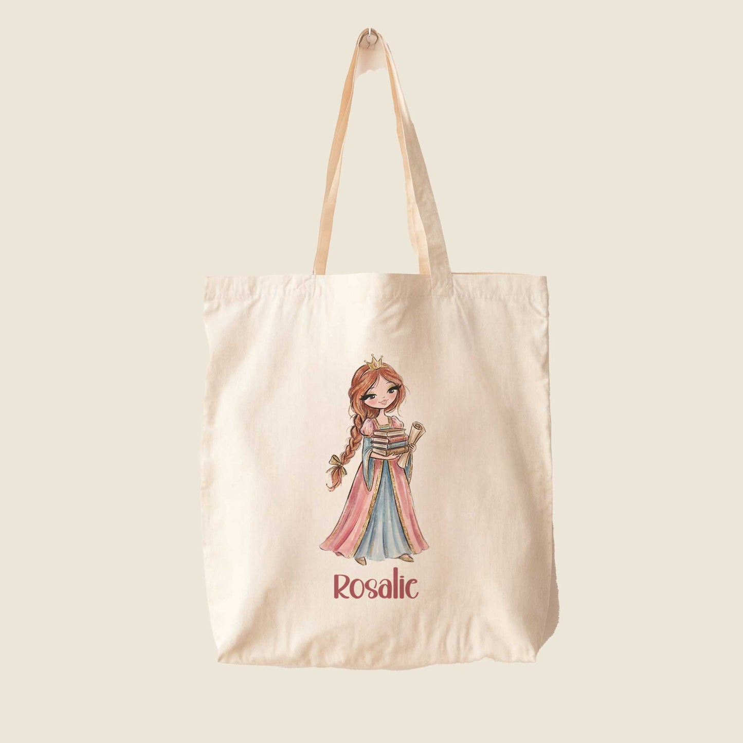 Beige totebag op een lichte effen achtergrond met een prinses die een stapel boeken en een opgerolde perkamentrol vasthoudt. Onder de illustratie staat de naam Rosalie.