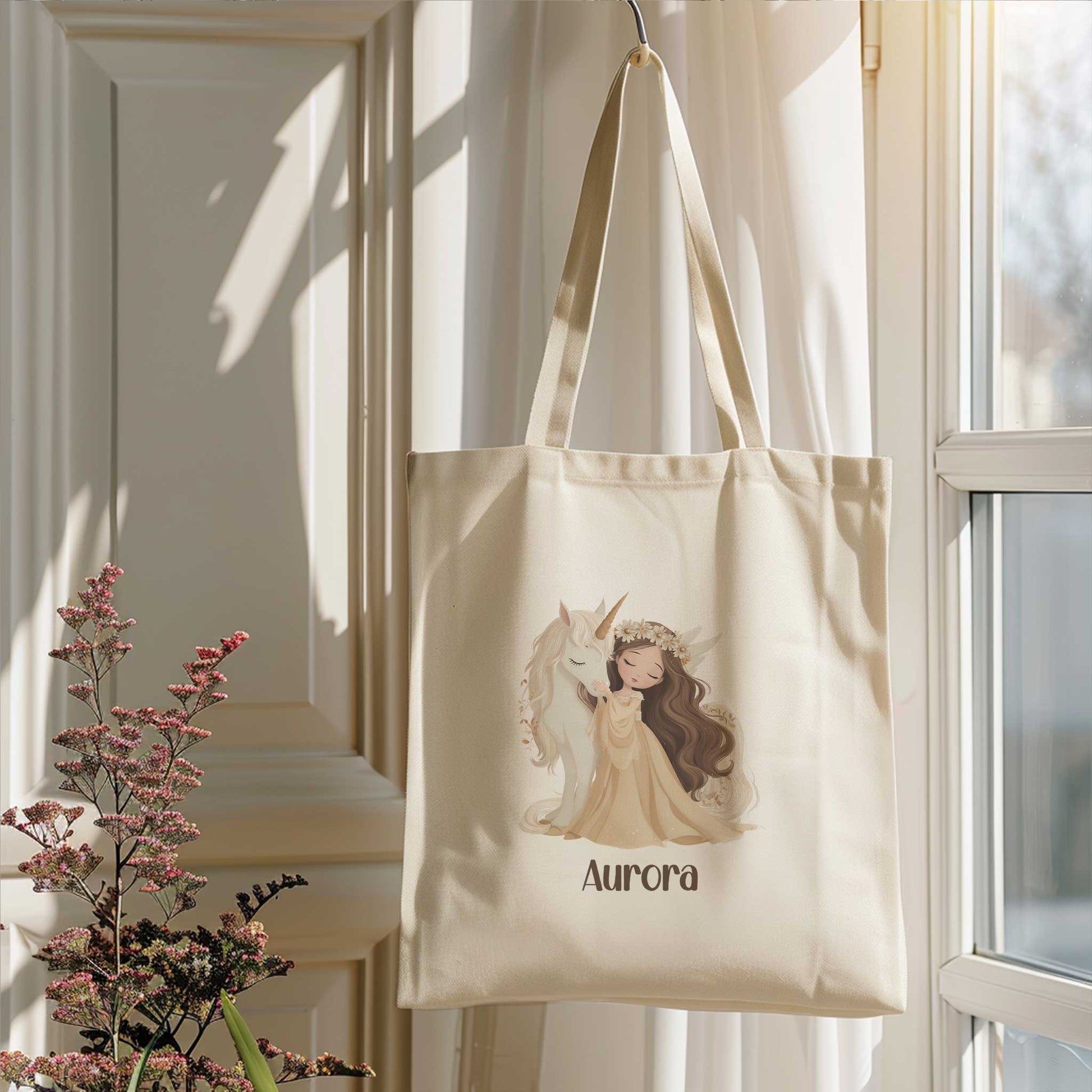 Beige totebag met een prinses die een witte eenhoorn knuffelt en de naam Aurora. De tas hangt voor een raam naast bloeiende takken.