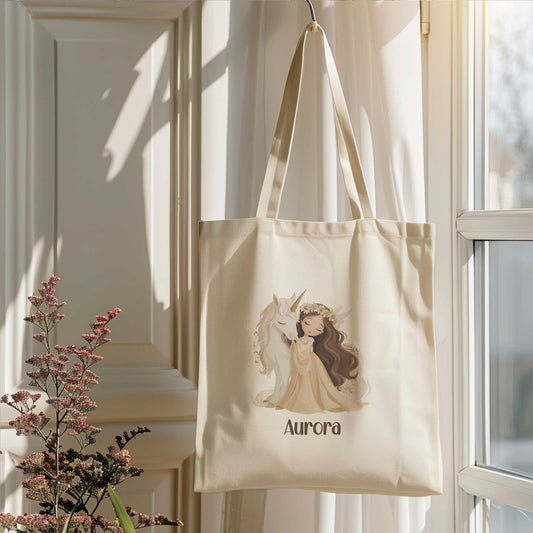 Beige totebag met een prinses die een witte eenhoorn knuffelt en de naam Aurora. De tas hangt voor een raam naast bloeiende takken.