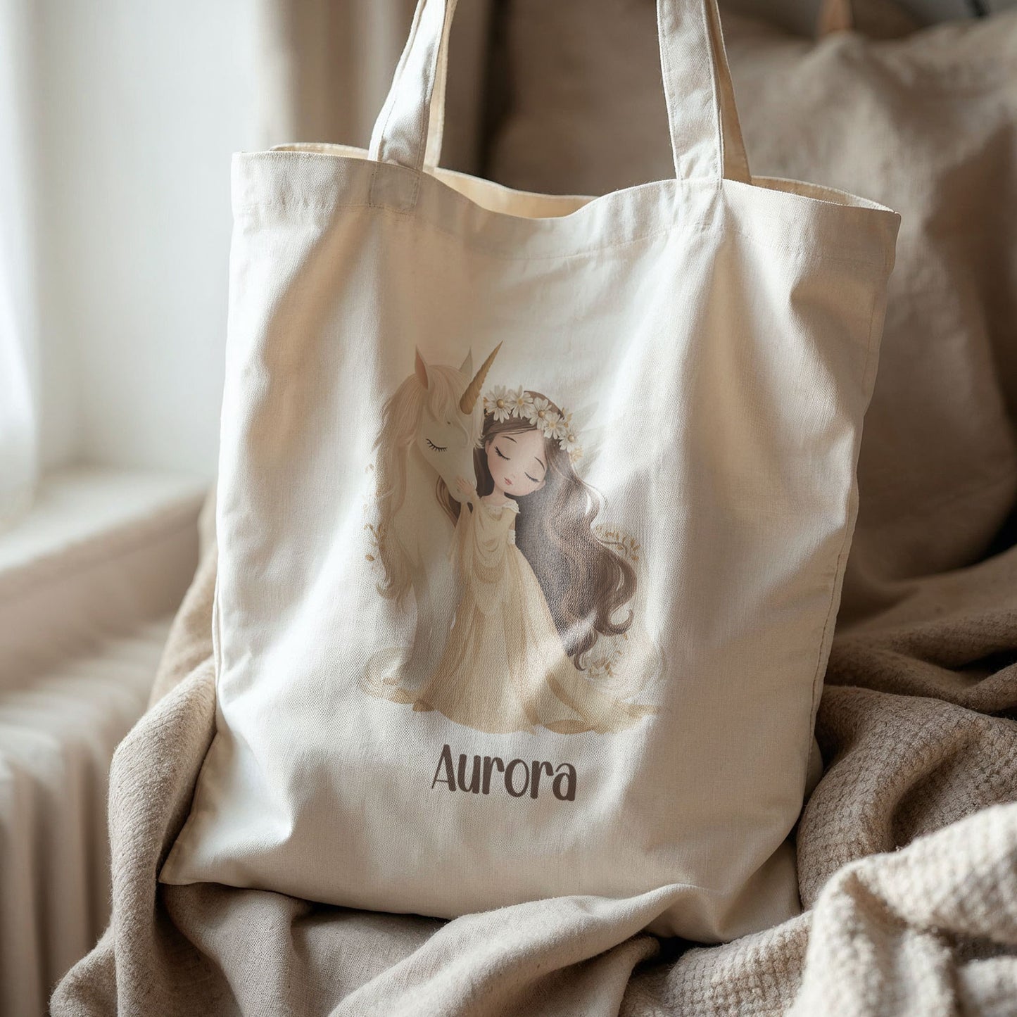 Beige totebag met een prinses en een witte eenhoorn met daaronder de naam Aurora. De tas ligt op een stoel met beige kussens en een deken.