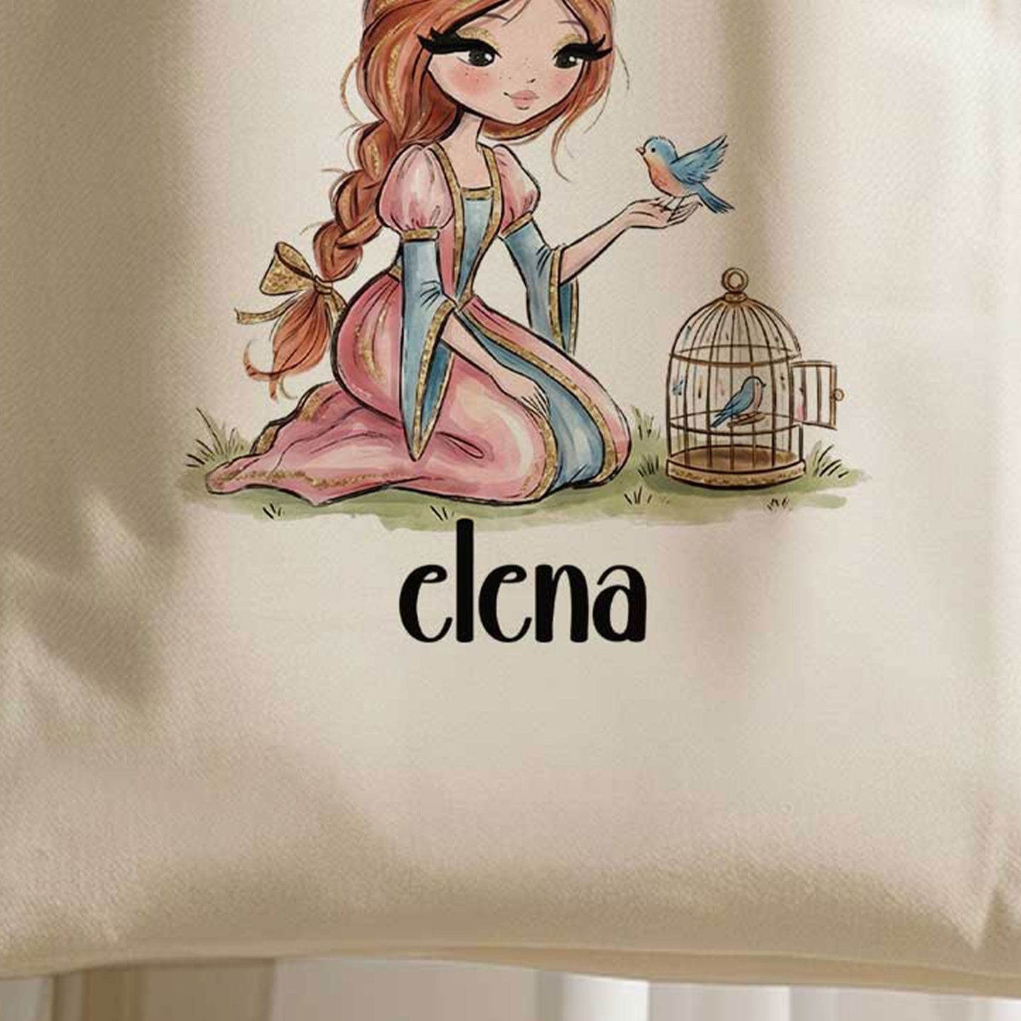 Close-up van een beige totebag met een illustratie van een knielende prinses met een vogeltje op haar hand en een vogelkooitje naast haar. Onder de illustratie staat de naam Elena