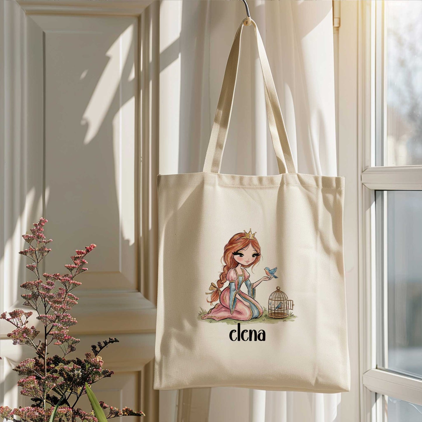 Beige totebag met een prinses, een blauw vogeltje en een vogelkooitje. Onder de illustratie staat de naam Elena. De tas hangt voor een raam naast bloeiende takken.