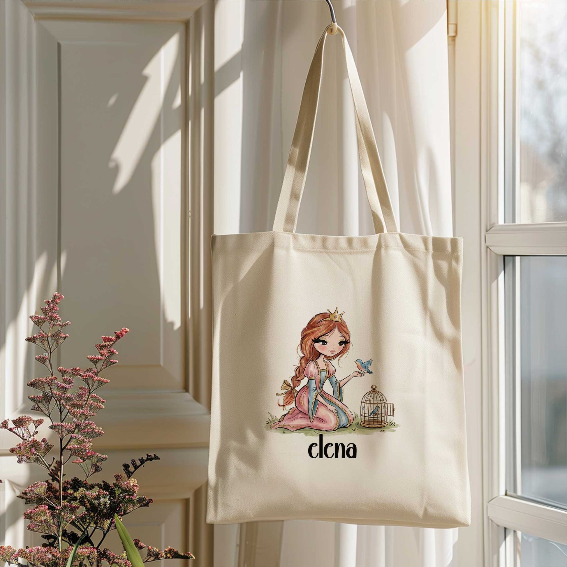 Beige totebag met een prinses, een blauw vogeltje en een vogelkooitje. Onder de illustratie staat de naam Elena. De tas hangt voor een raam naast bloeiende takken.