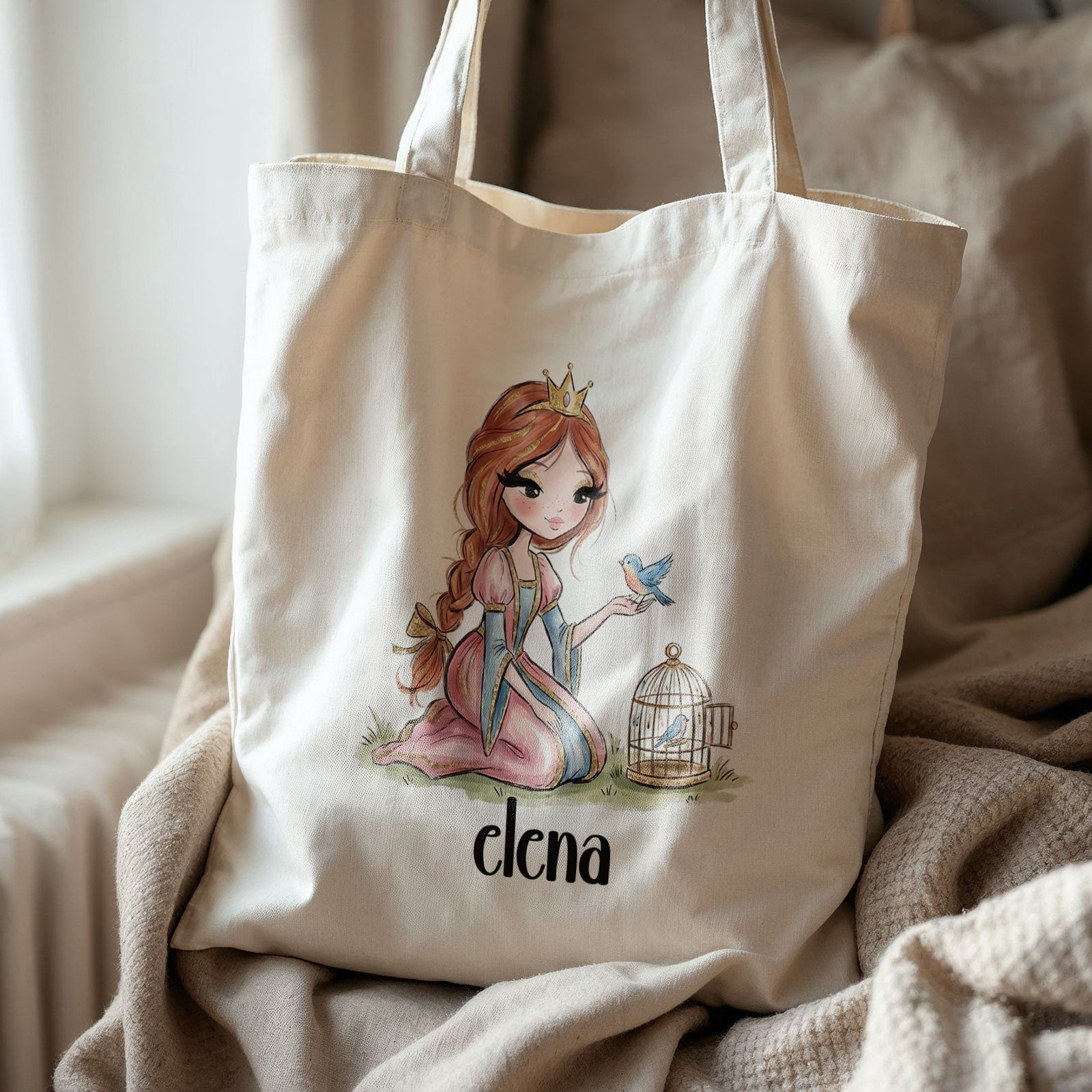 Beige totebag met een prinses die een blauw vogeltje op haar hand heeft en een vogelkooitje naast zich. Onder de illustratie staat de naam Elena. De tas ligt op een stoel met beige kussens en een deken.