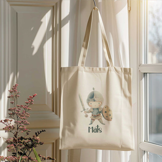 Crèmekleurige katoenen totebag met een ridder met zwaard en schild en de naam Mats, hangend naast een raam in het zonlicht.