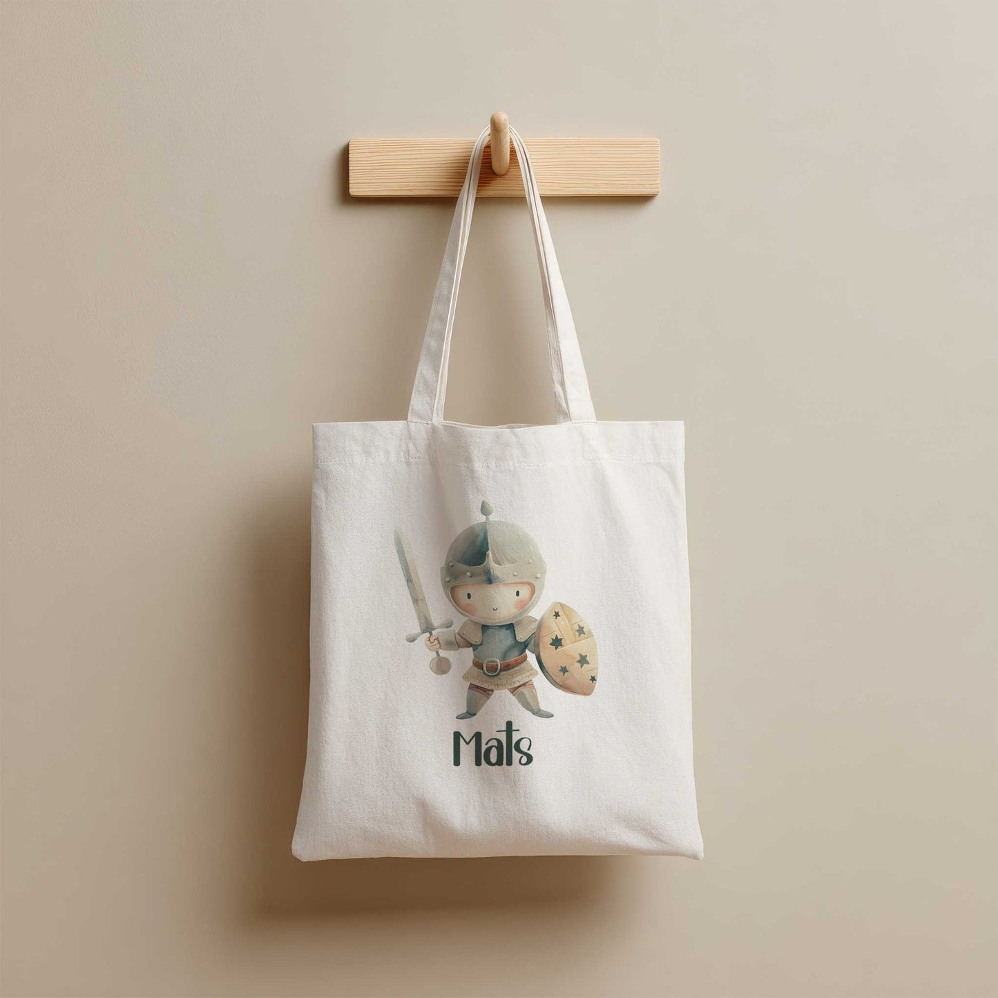 Crèmekleurige katoenen totebag met een illustratie van een ridder met zwaard en schild en de naam Mats, hangend aan een houten kapstok.