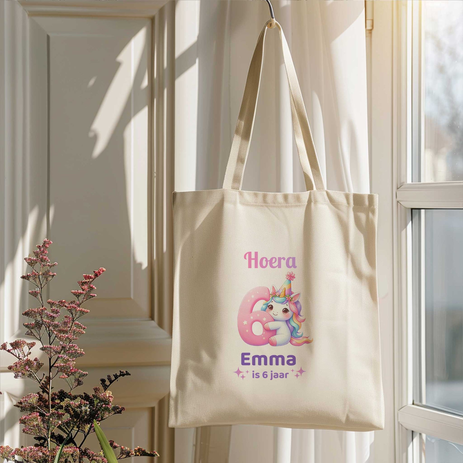 Lichte totebag hangt aan een haak bij een raam. Op de tas staat een unicorn, een groot roze cijfer 6 en de tekst Hoera Emma is 6 jaar.