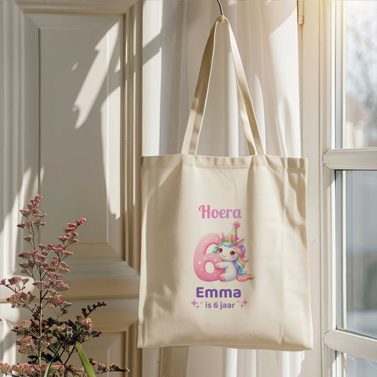Lichte totebag hangt aan een haak bij een raam. Op de tas staat een unicorn, een groot roze cijfer 6 en de tekst Hoera Emma is 6 jaar.