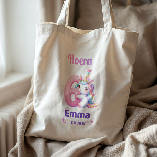 Katoenen totebag met unicorn-illustratie en tekst ‘Hoera Emma is 6 jaar’ op bed met beige plaid