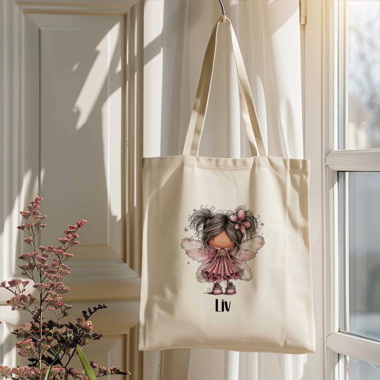 Beige totebag hangend bij een raam, bedrukt met een fee met roze vleugels en de naam Liv.