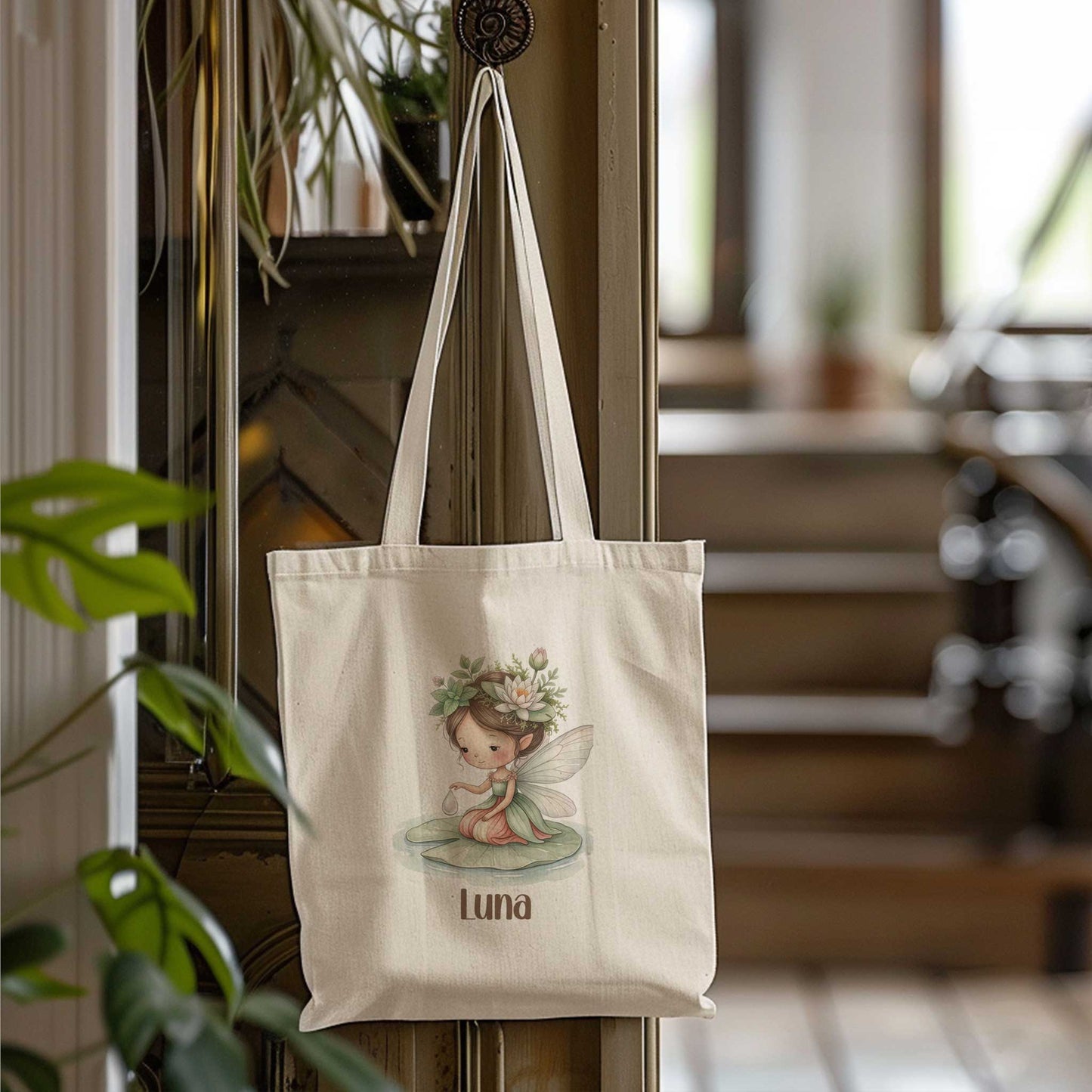 Beige totebag die aan een haak hangt in een huiselijke ruimte, met een illustratie van een fee op een waterlelieblad en de naam Luna.