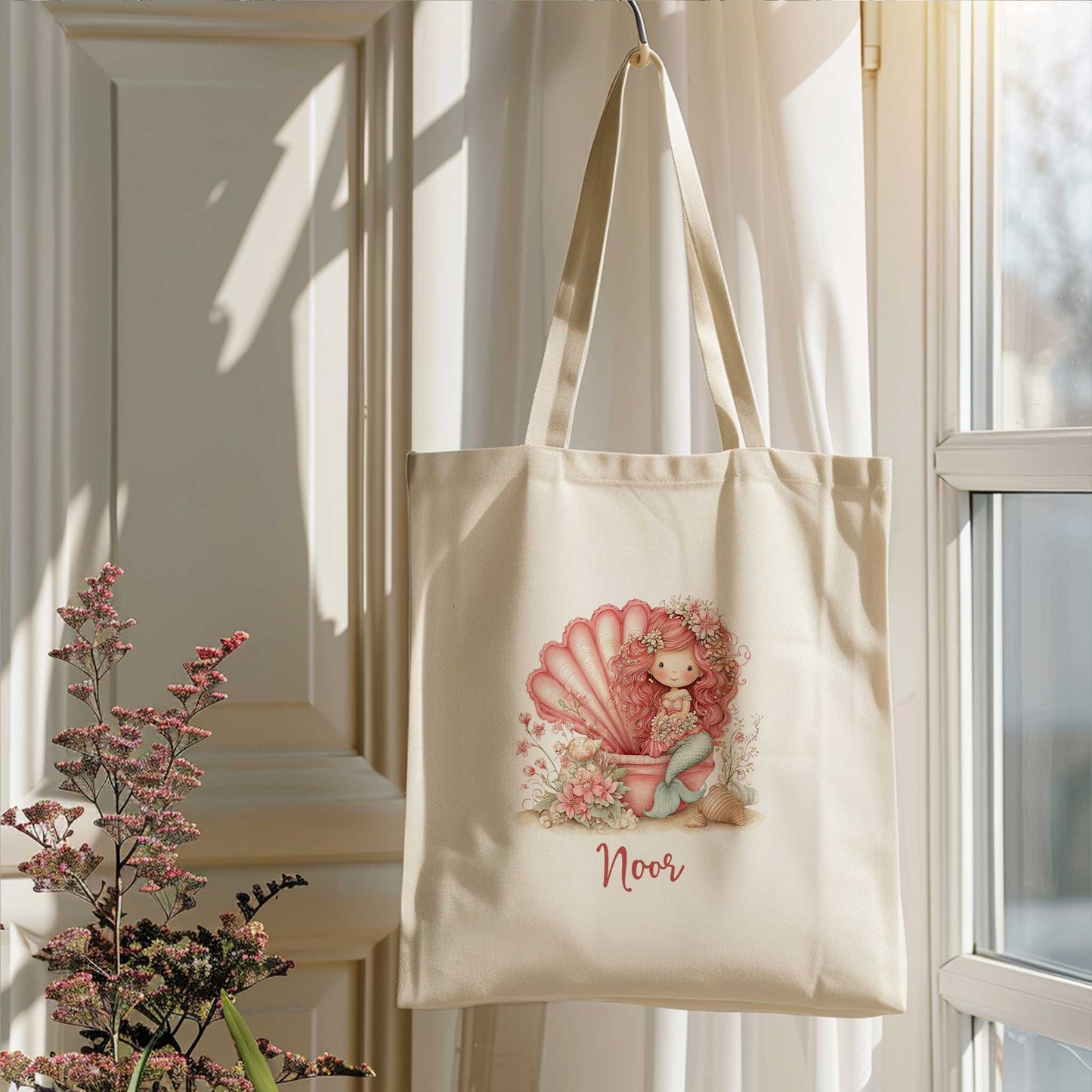 Gepersonaliseerde totebag met zeemeerminontwerp hangend bij een raam. De illustratie toont een roze zeemeermin in een grote schelp met bloemen en schelpen. Onder de afbeelding staat de naam Noor.