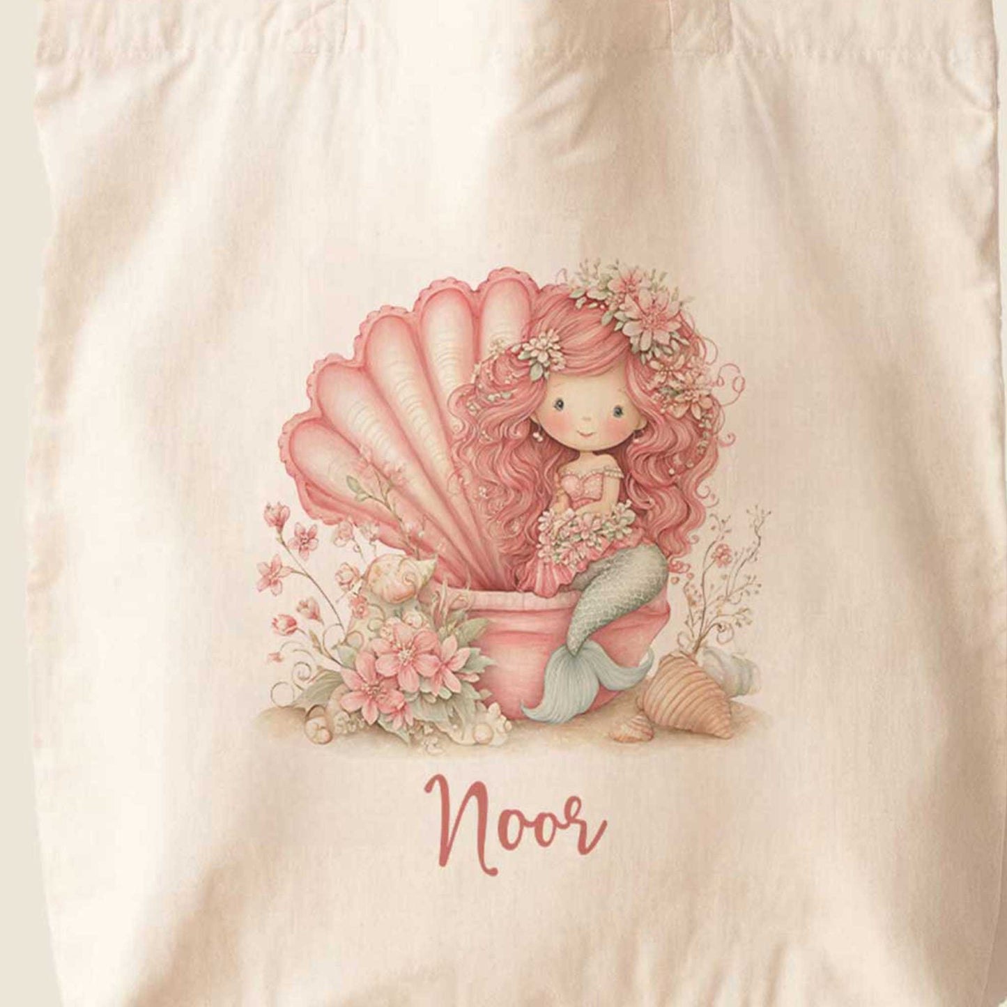 Close-up van een totebag met een gedetailleerde illustratie van een roze zeemeermin in een open schelp. Rondom de zeemeermin staan roze bloemen, kleine schelpjes en zachte zeedetails. De naam Noor staat onder het ontwerp.