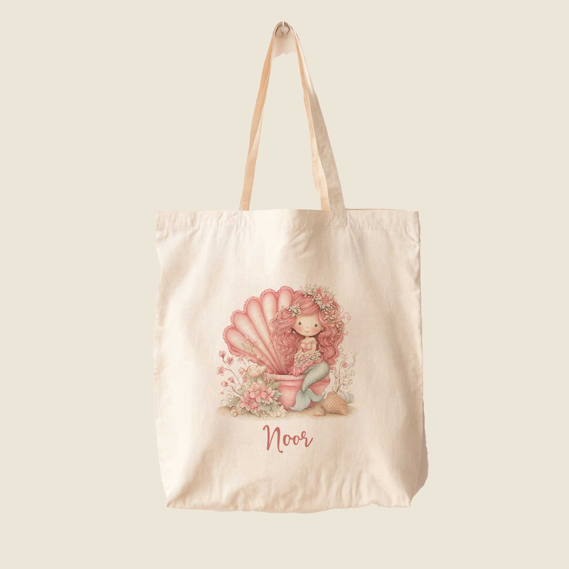 Vrijstaande katoenen totebag op een lichte achtergrond met een gepersonaliseerde zeemeerminillustratie. De roze zeemeermin zit in een grote schelp tussen bloemen en schelpen met daaronder de naam Noor