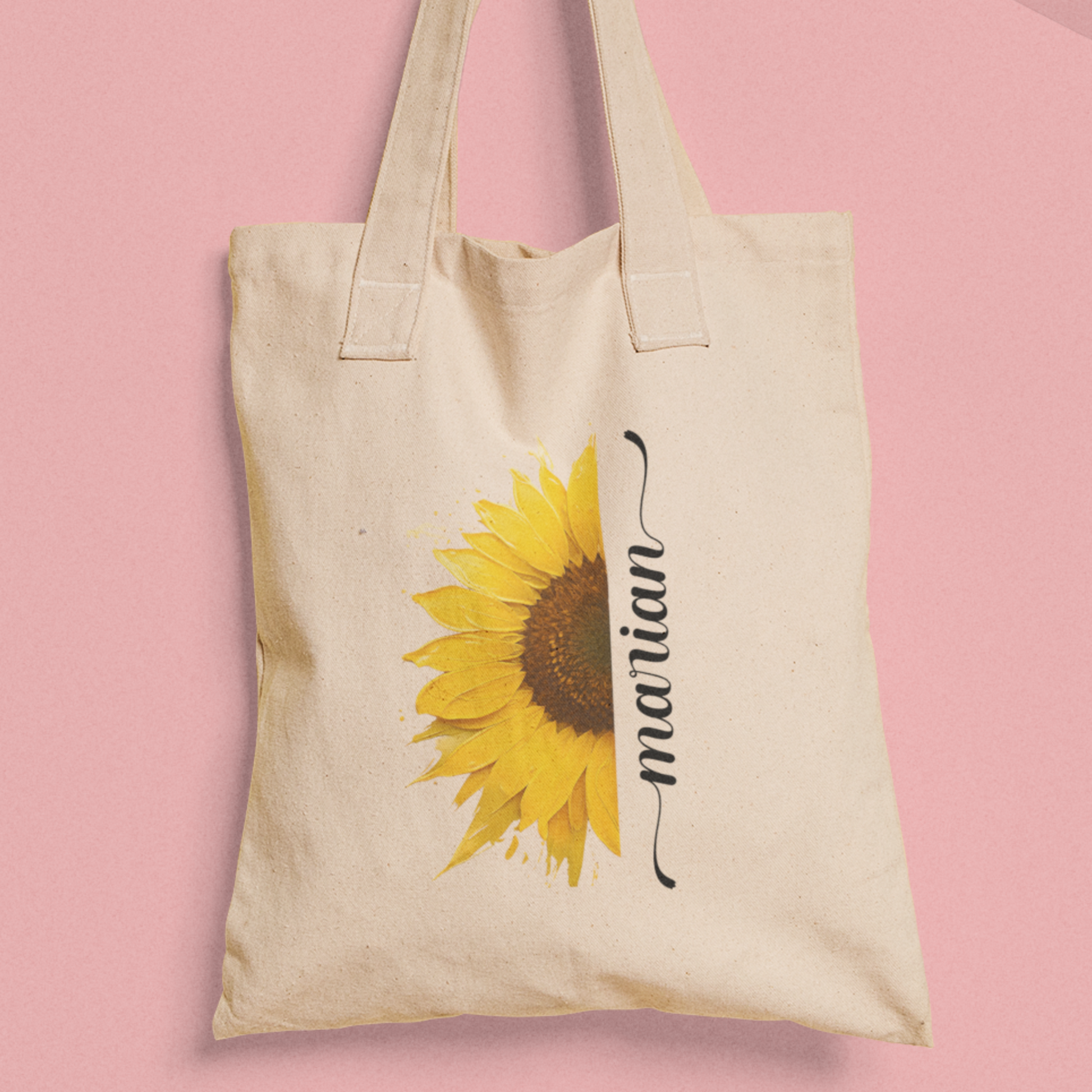 Totebag met zonnebloem en naam Marian, tegen roze achtergrond.