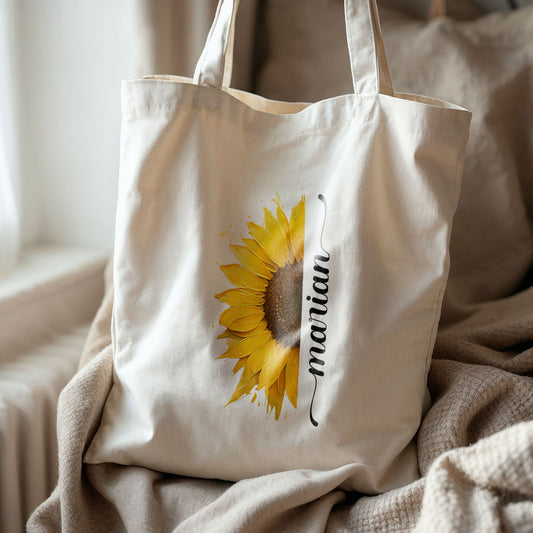 Totebag van katoen met grote zonnebloem en naam Marian, gefotografeerd op een stoel bij raam met dekens.