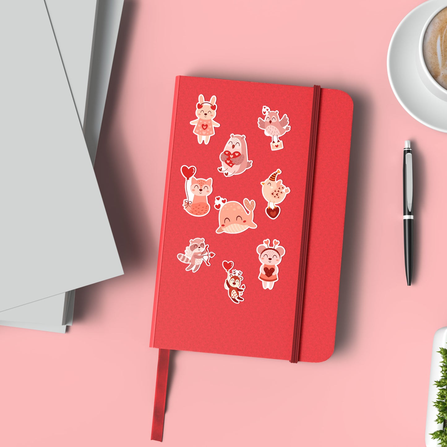 Rood notitieboek op roze ondergrond met verschillende valentijn dierenstickers, naast een kop koffie en pen.