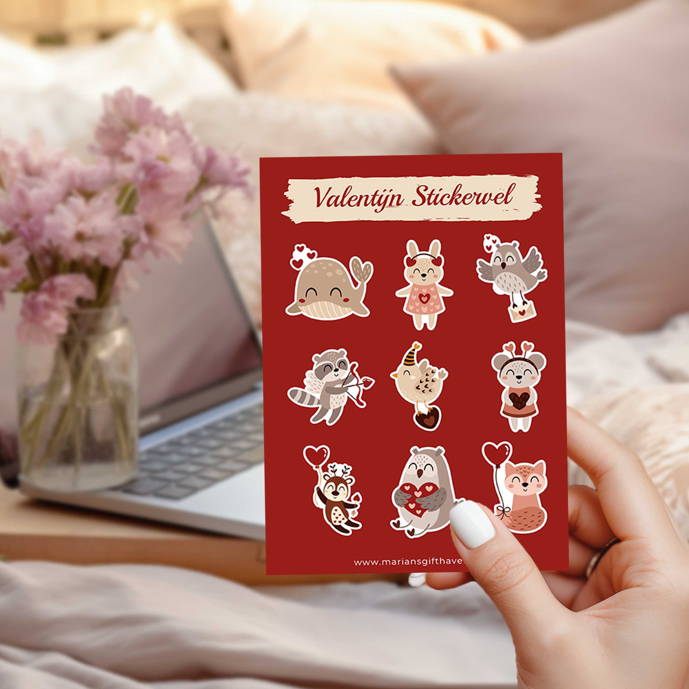 Hand houdt een rood valentijn stickervel met schattige dierenillustraties en hartjes, met laptop en bloemen op de achtergrond.