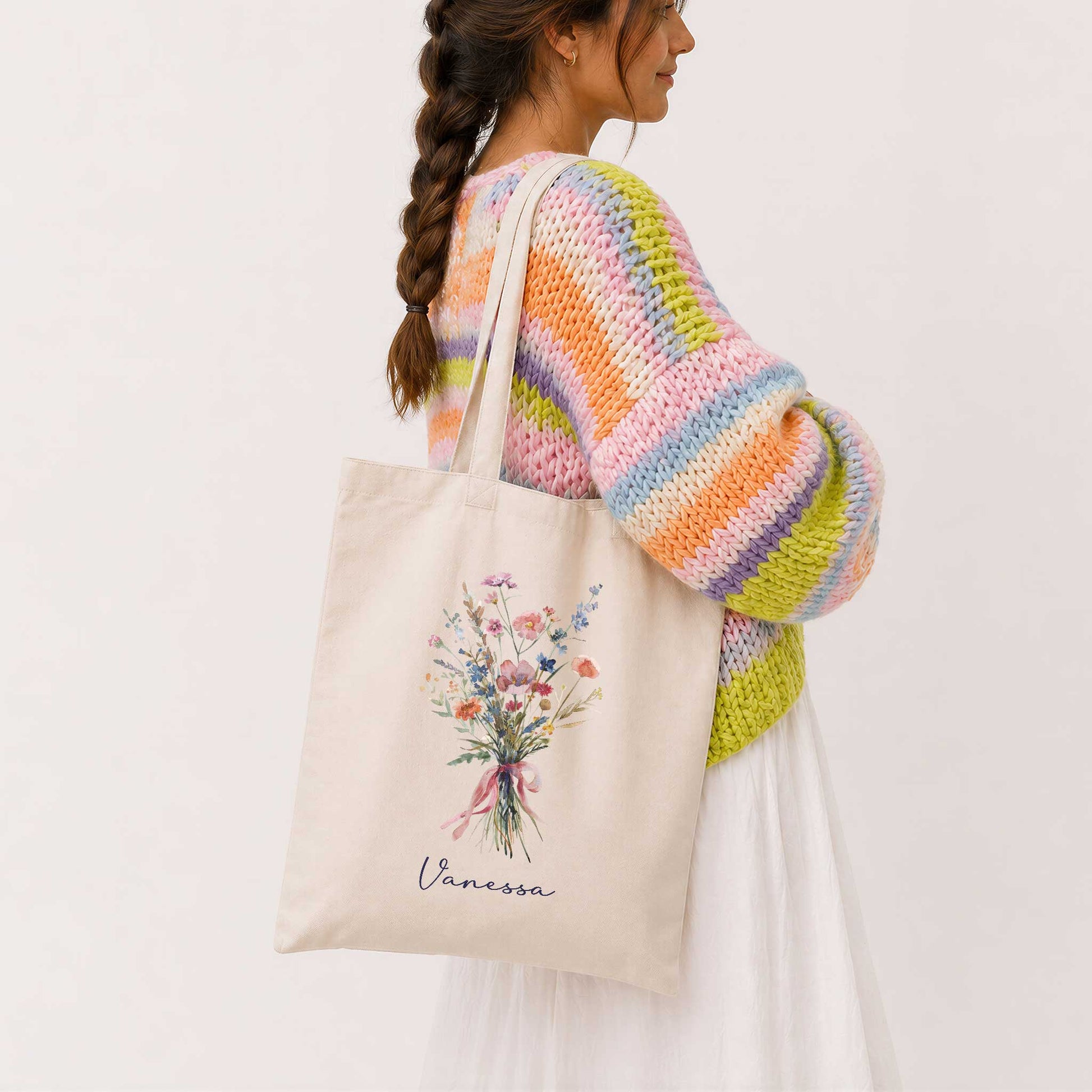 Vrouw met lange vlecht draagt een beige totebag met een boeket wilde bloemen in zachte pastelkleuren en de naam Vanessa. Ze draagt een kleurrijke gebreide trui in roze, oranje, groen en paars tinten.