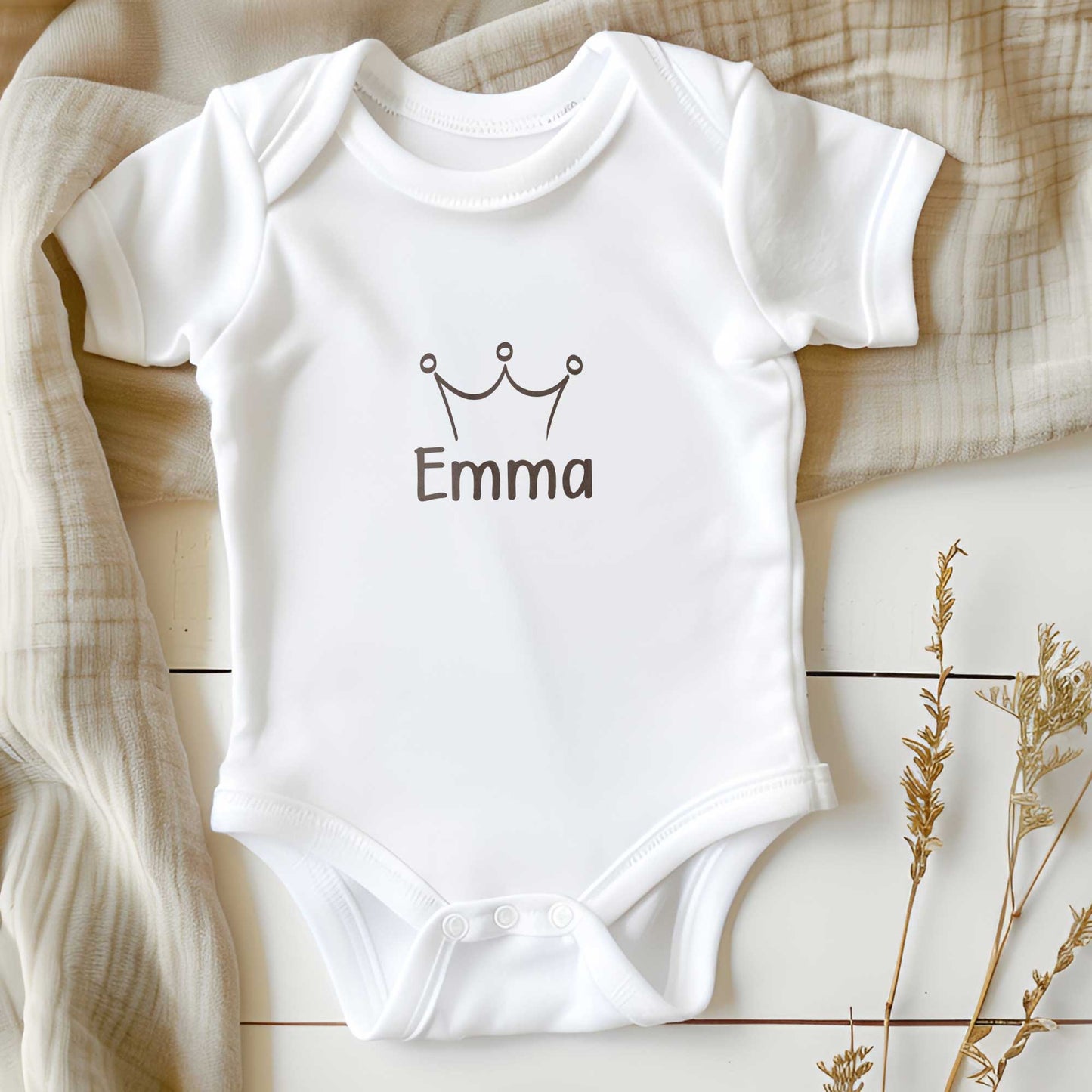 Wit babyrompertje met gepersonaliseerde naam Emma en taupekleurig kroontje liggend op een zachte neutrale achtergrond.