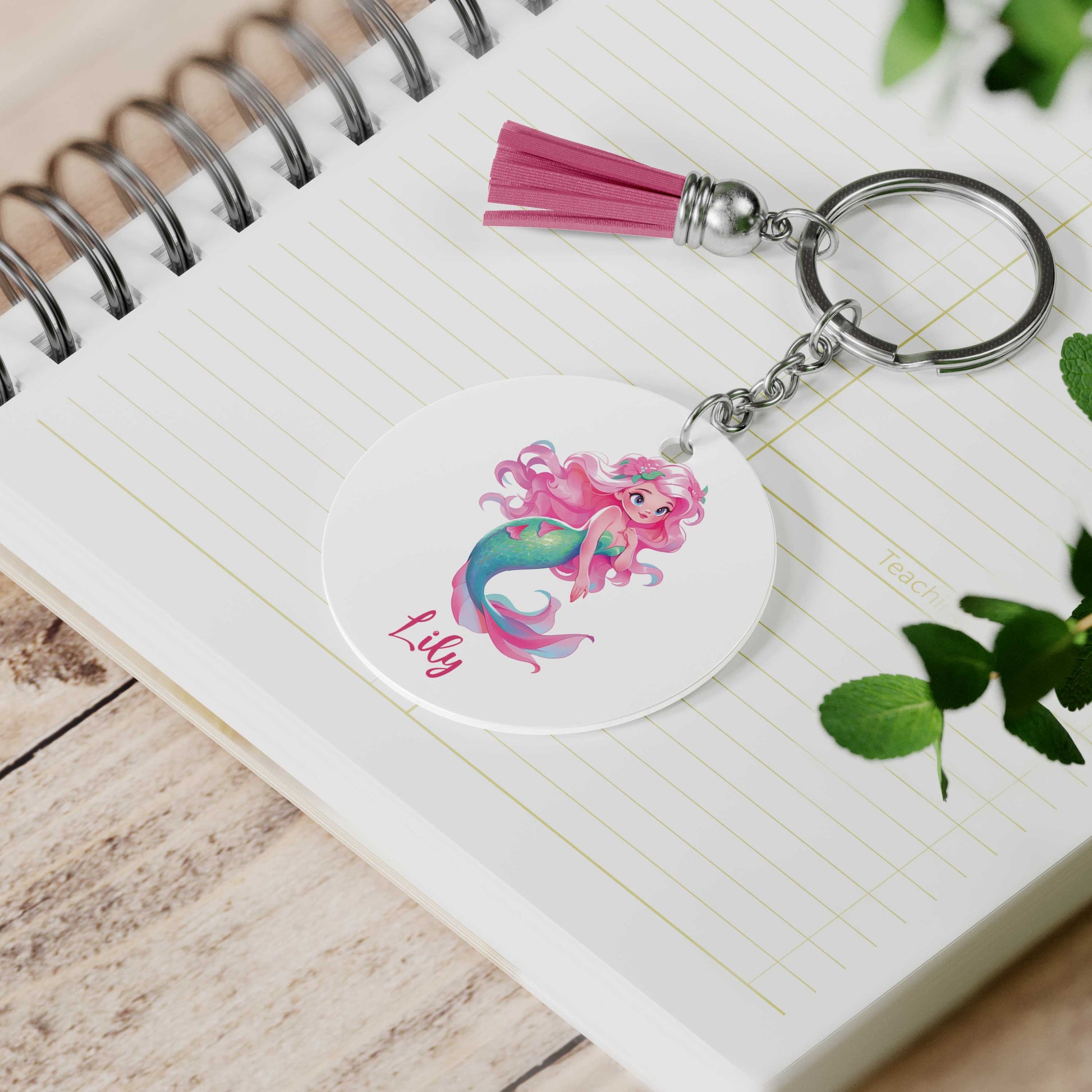 Gepersonaliseerde sleutelhanger met roze kwastje liggend op een open notitieboek. Op de hanger staat een zeemeermin met bloemenhaar en de naam Lily.