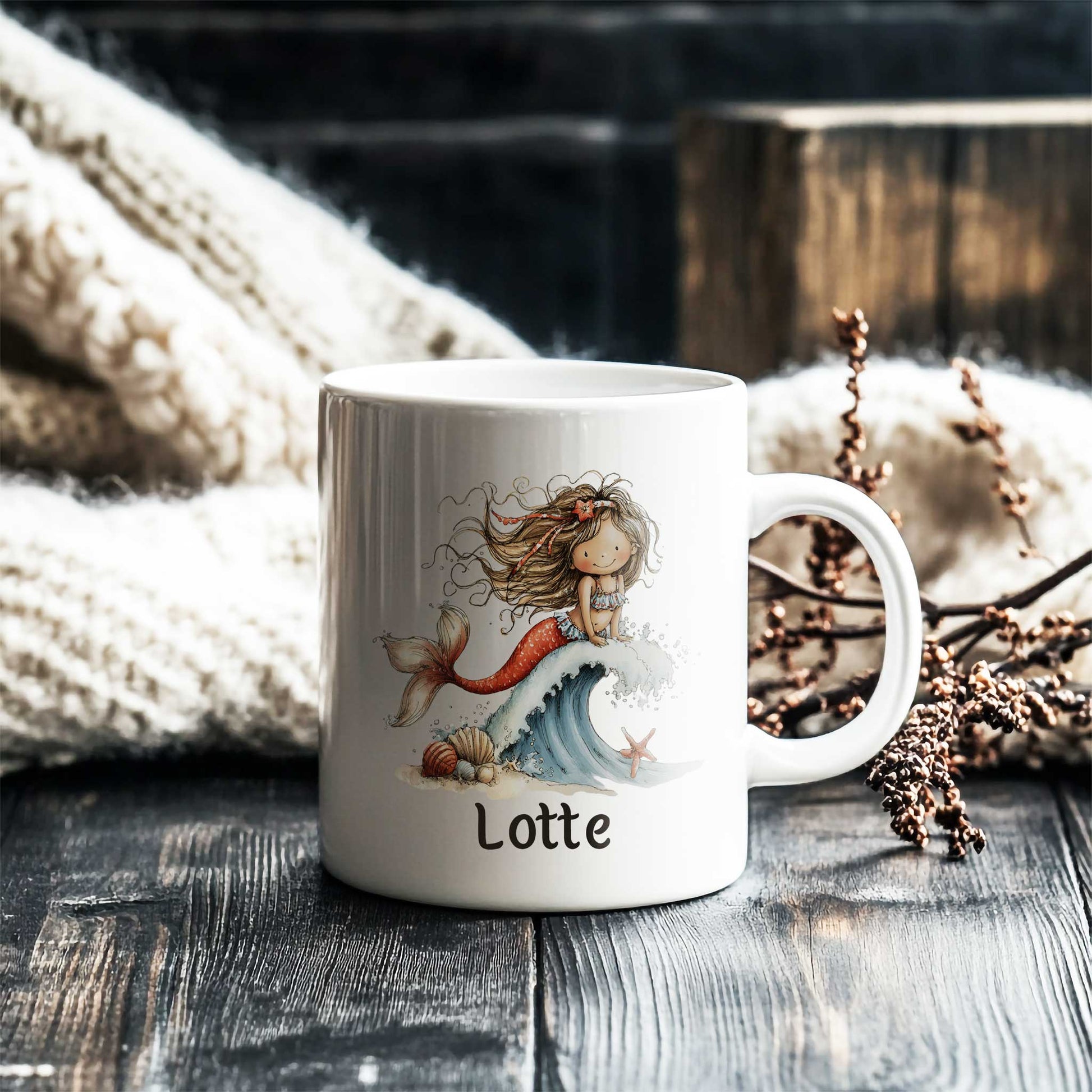 Witte mok met een zeemeermin op een golf en de naam Lotte op een donkere houten tafel naast een gebreide deken en decoratieve takken.