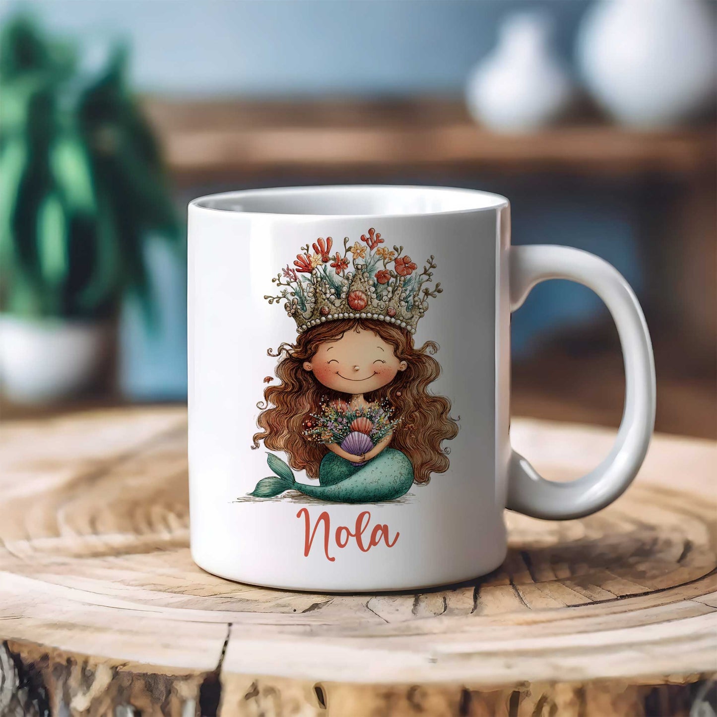 Witte mok met zeemeerminillustratie en naam Nola op een houten tafel met een onscherpe achtergrond van planten en woondecoratie.