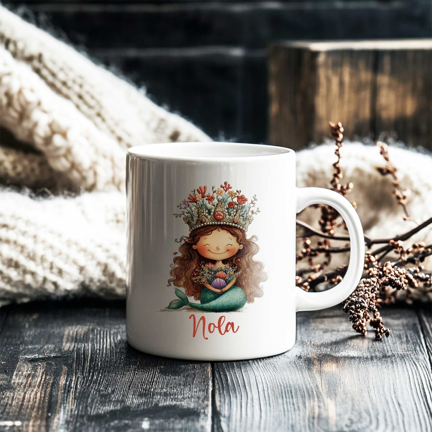 Witte mok met zeemeermin en naam Nola op een donkere houten tafel naast een gebreide deken en decoratieve takken.