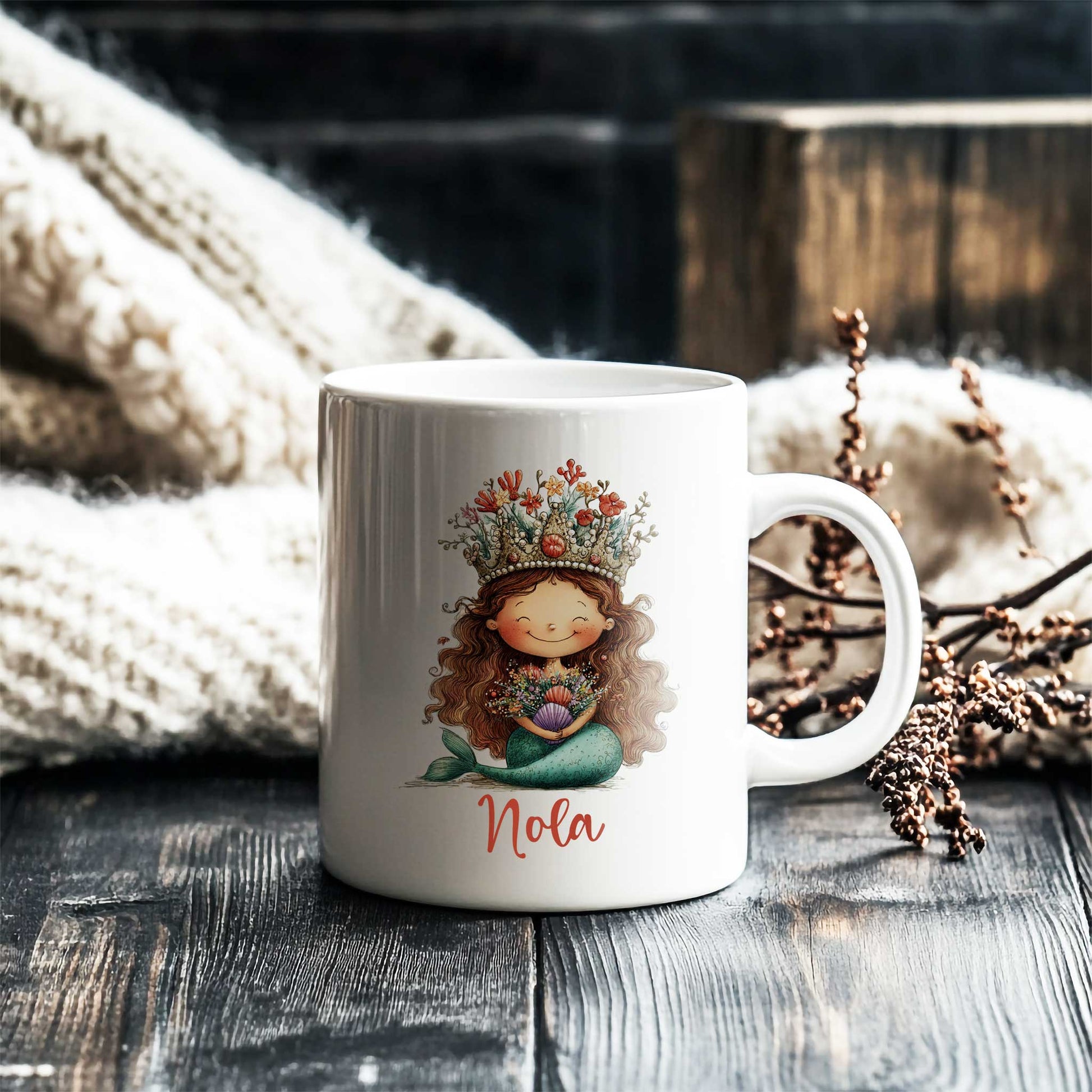Witte mok met zeemeermin en naam Nola op een donkere houten tafel naast een gebreide deken en decoratieve takken.