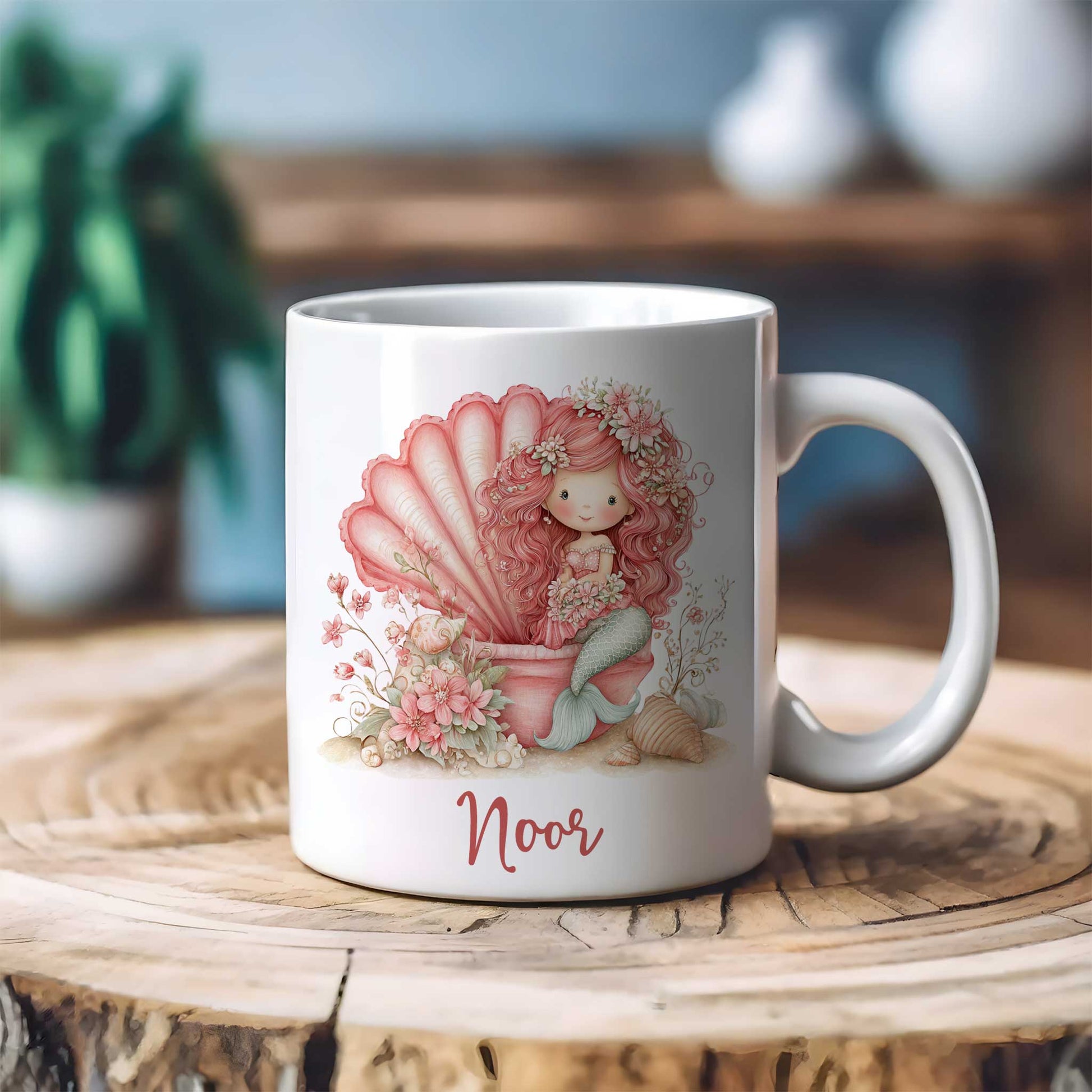 Witte keramische mok met gepersonaliseerde naam Noor op een houten tafel. Op de mok staat een roze zeemeermin die in een grote schelp zit, omringd door bloemen en schelpen in zachte pastelkleuren.