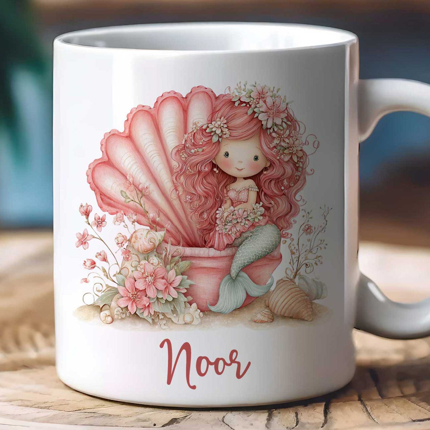 Close-up van een witte mok met een gedetailleerde illustratie van een roze zeemeermin. De afbeelding laat de grote schelp, bloemen, schelpen, mintgroene staart en de naam Noor duidelijk zien.