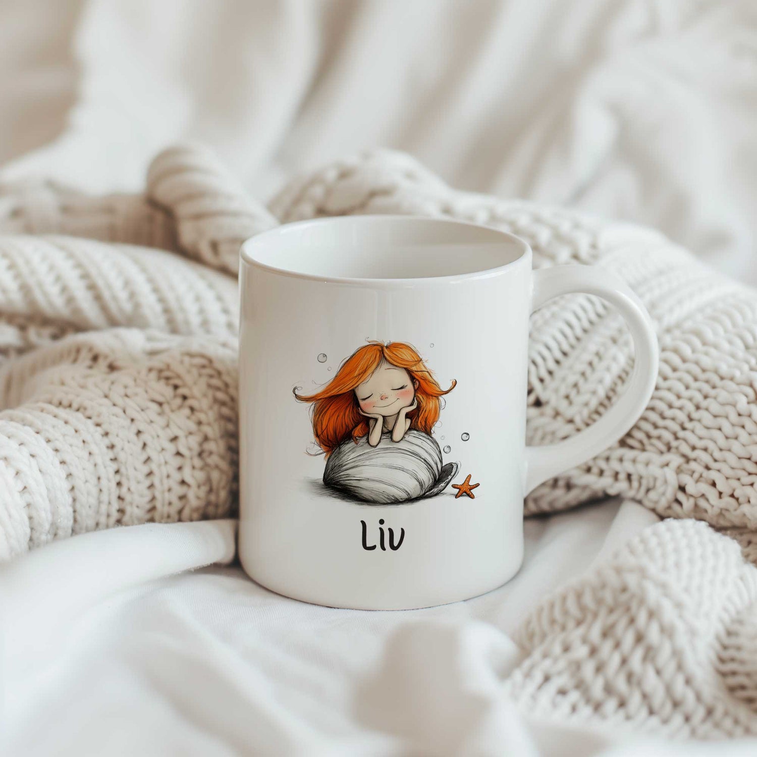 Witte keramische mok met een illustratie van een dromende zeemeermin met lang oranje haar op een grote witte schelp. Onder de illustratie staat de naam Liv. op gebreide achtergrond